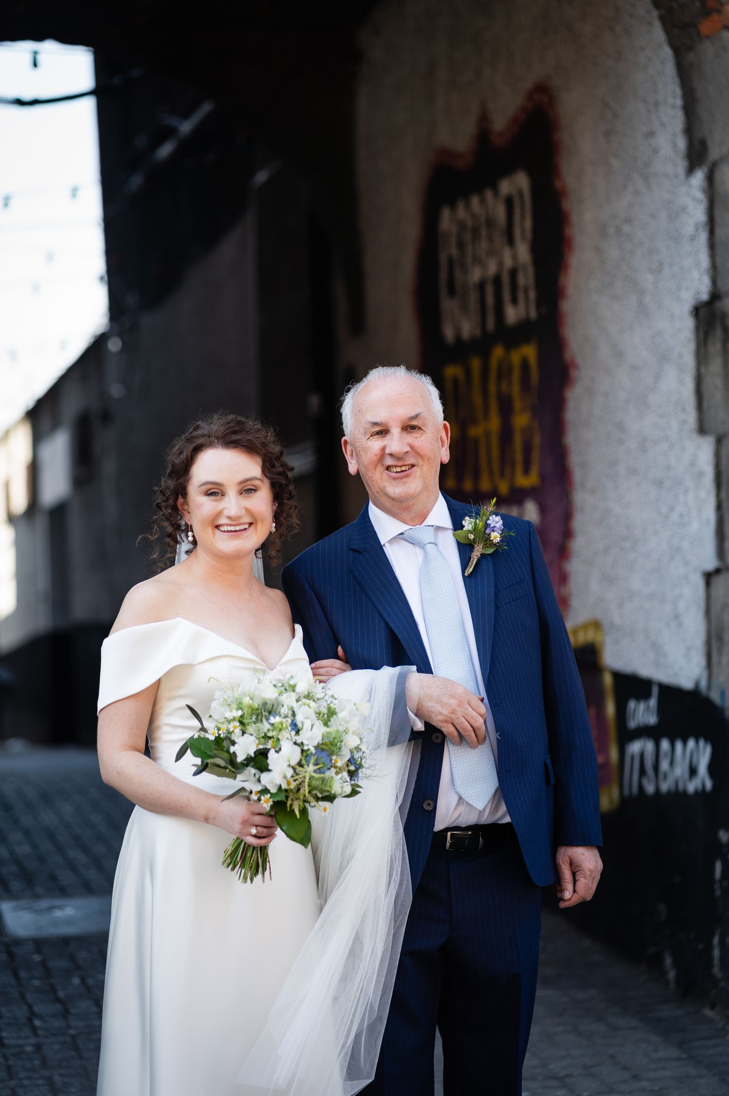 A heartwarming wedding photo of a pair in a white gown and navy suit standing together against an urban wall.