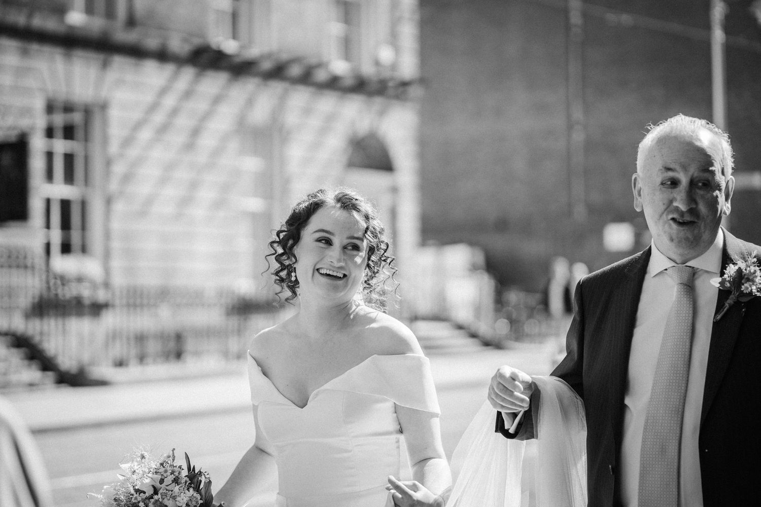 A radiant bride in an off-shoulder wedding dress walks arm in arm with a gentleman down an elegant hallway.
