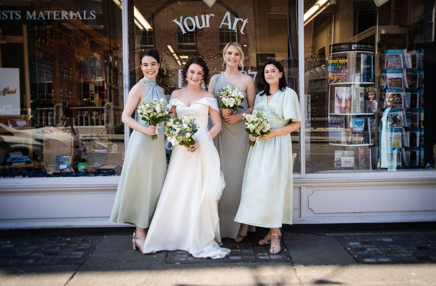 A bridal party poses in mint green and white dresses outside an art supply store storefront.