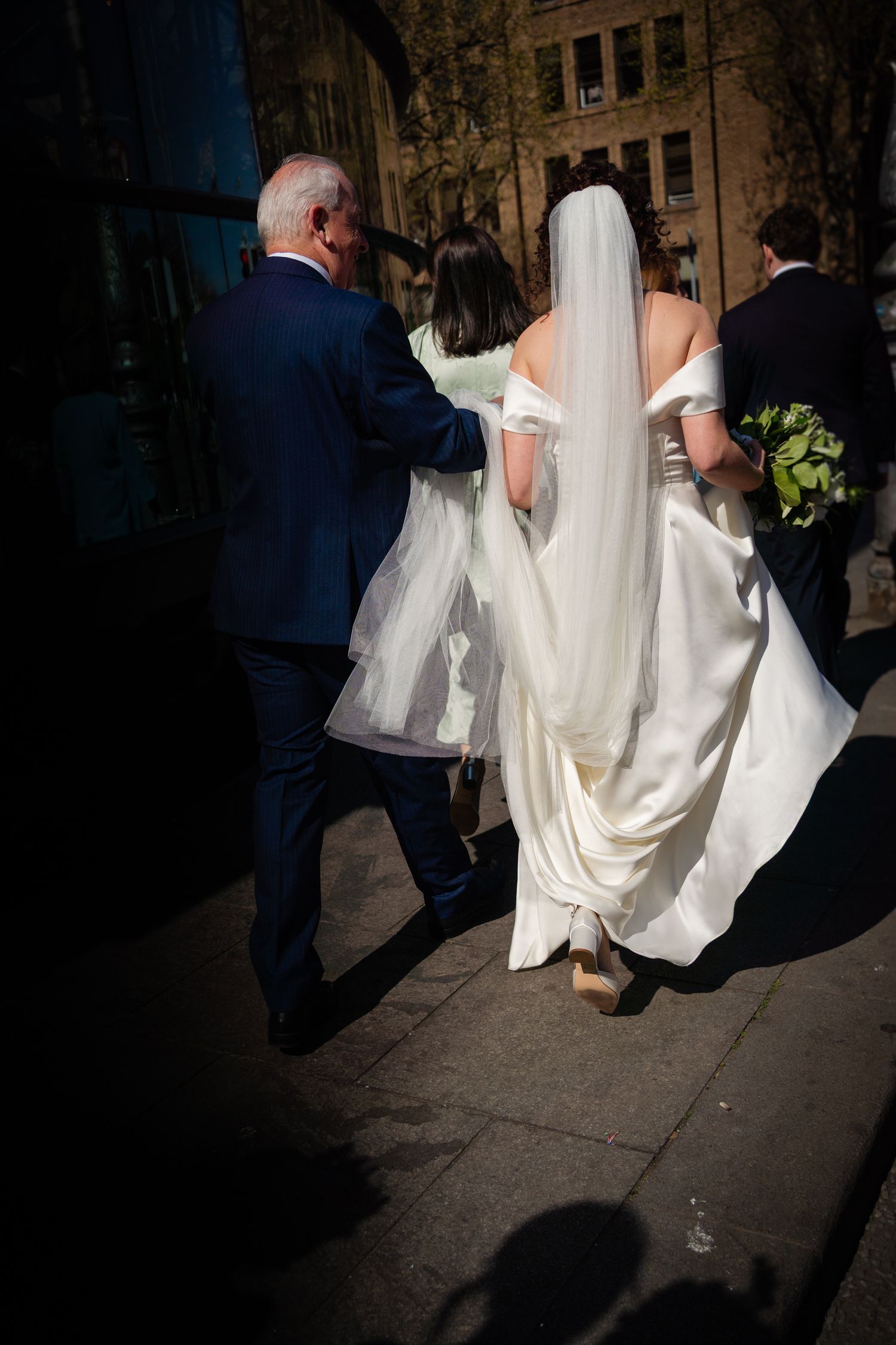 Walking scene of a bride in a flowing white dress and veil with other wedding party members on a city sidewalk.