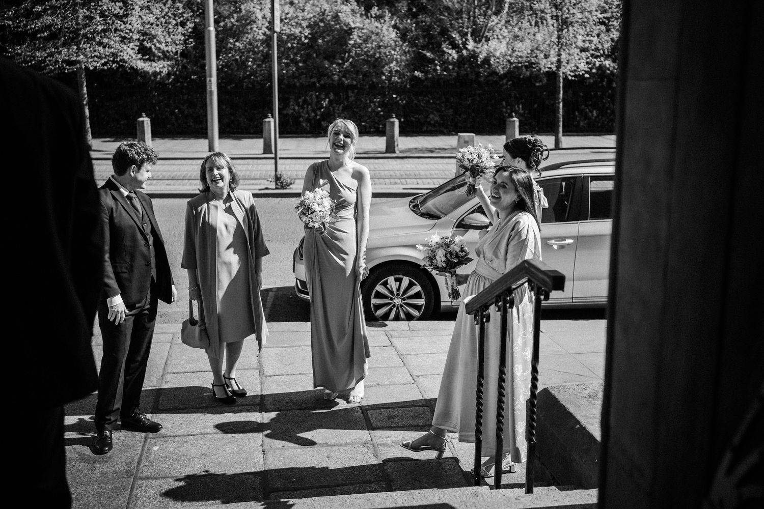 Black and white photo of a group gathering outdoors by a car in bright sunlight.