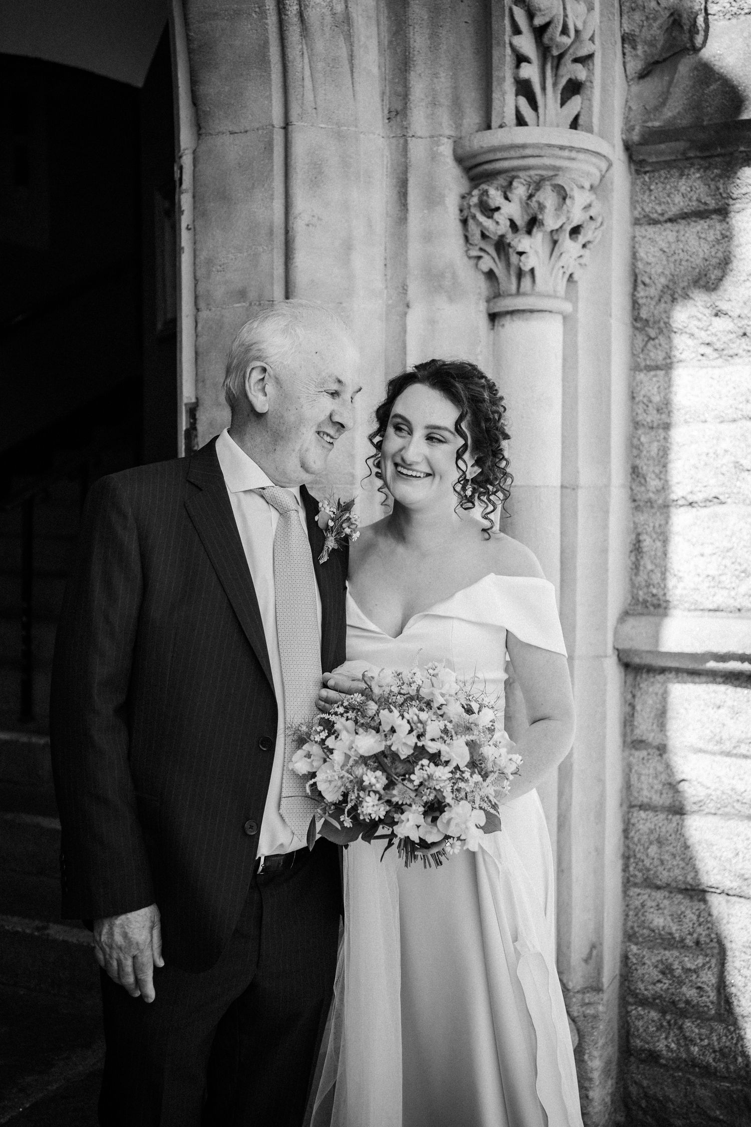 Black and white wedding photo showing an older gentleman and woman in formal attire standing by ornate stone pillar.