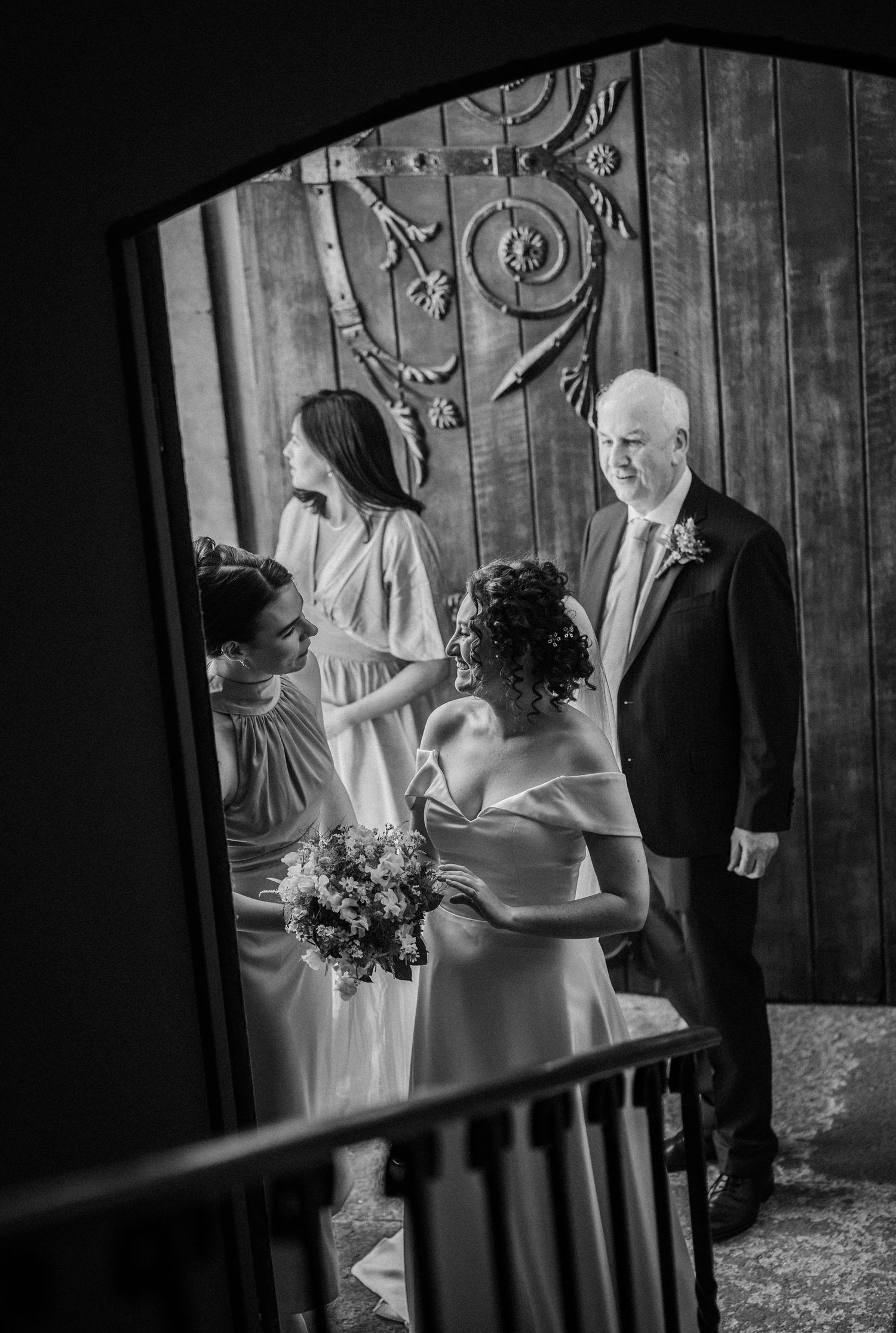 A black and white wedding photograph shows a group on elegant stairs with wooden doors and decorative metalwork in the background.