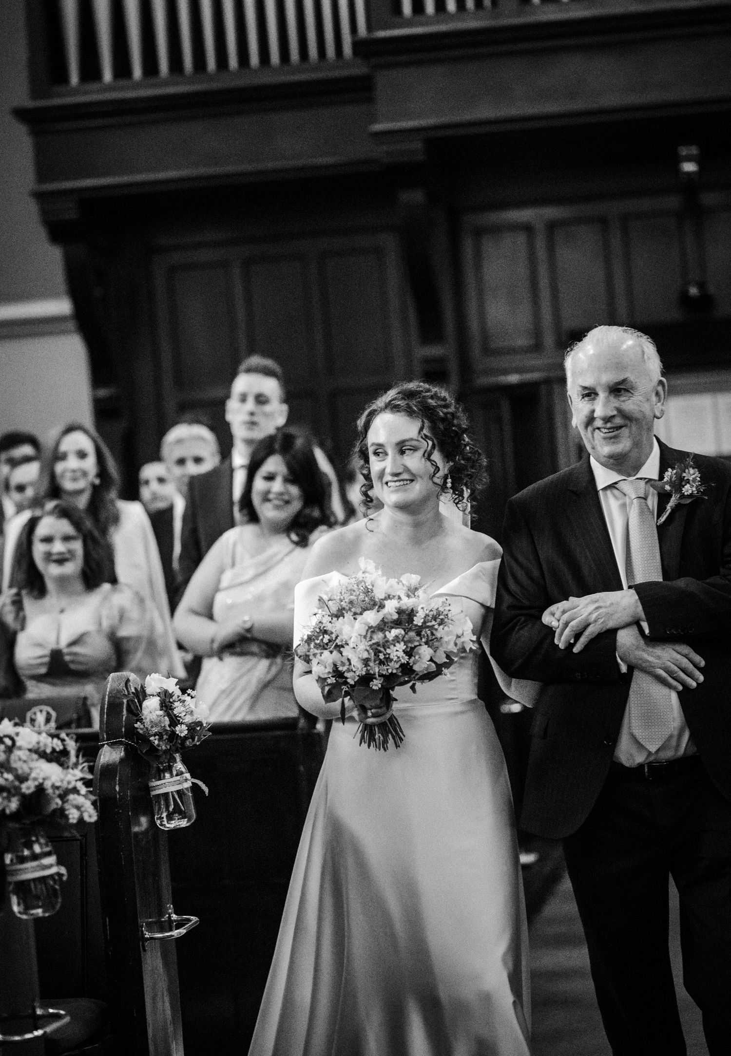 Black and white wedding photograph shows a joyful procession walking down a church aisle with flowers and decorative lighting.