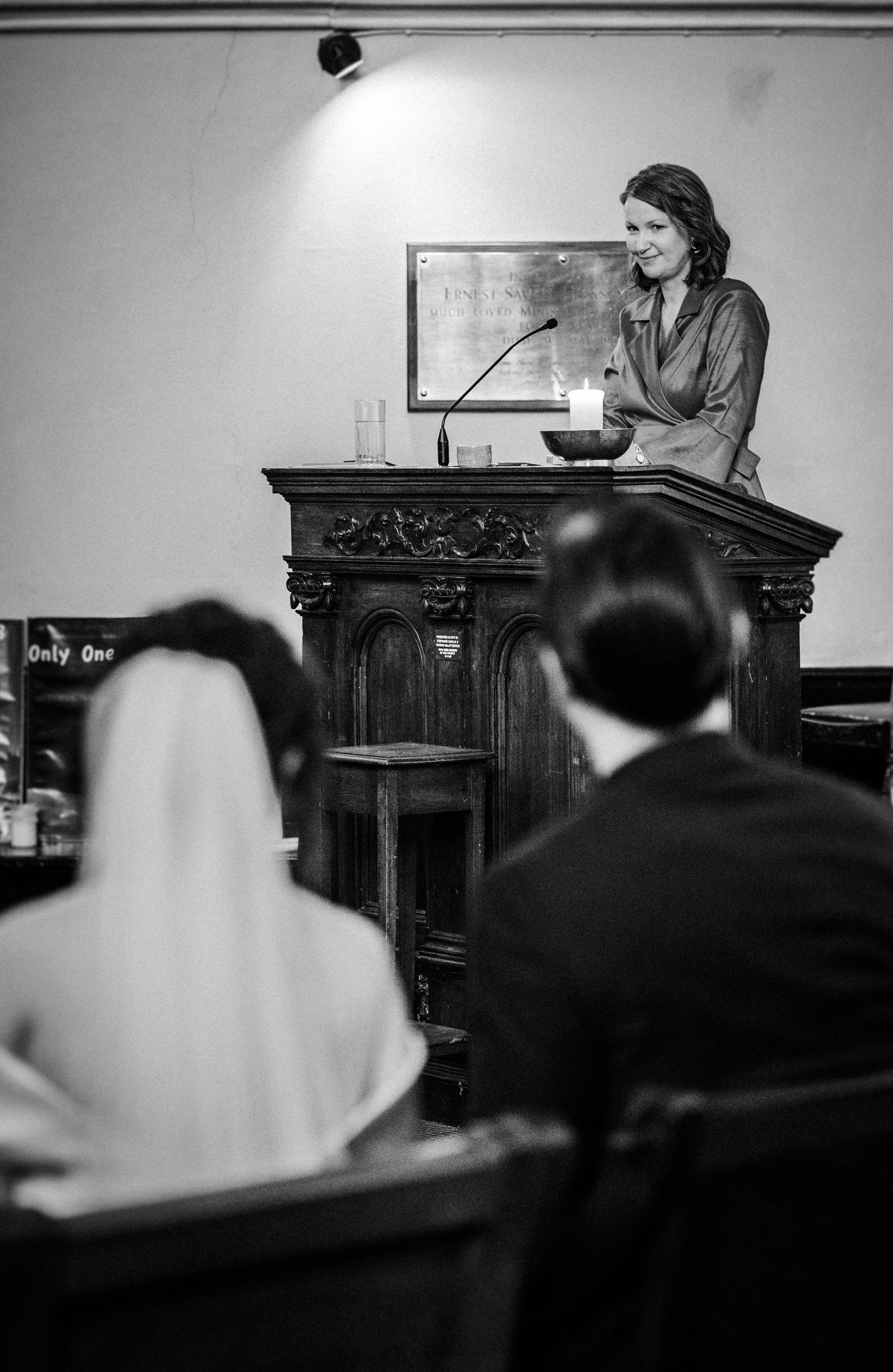 A black and white photograph shows a speaker at an ornate wooden podium addressing an audience in a formal setting.