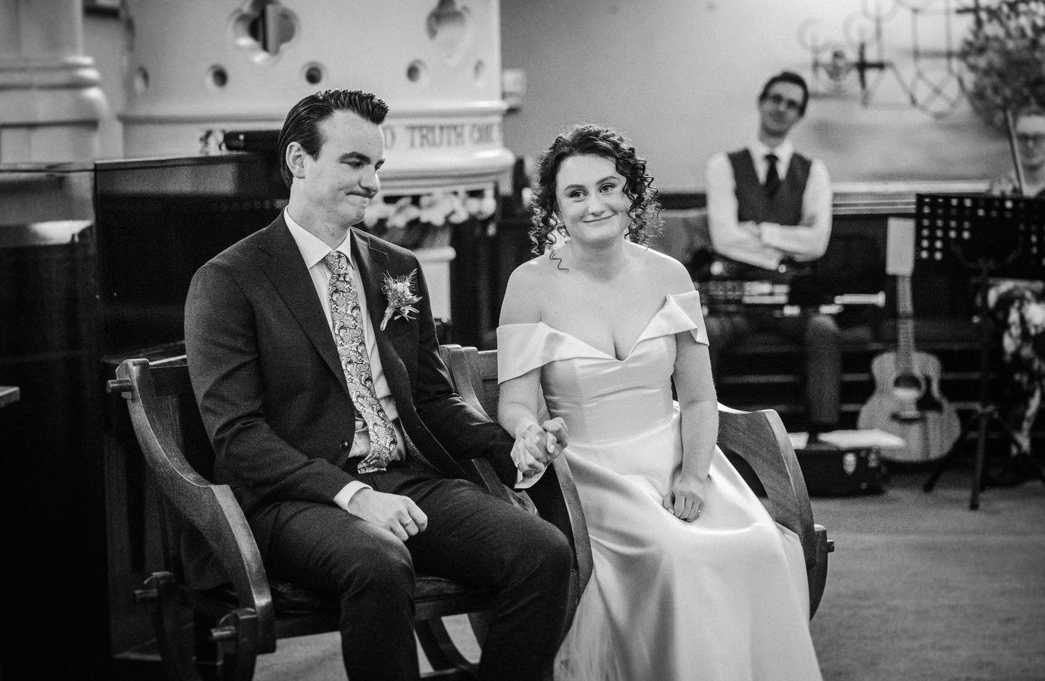 A vintage black and white photo shows a couple sitting together in formal attire at an indoor venue with someone standing behind.