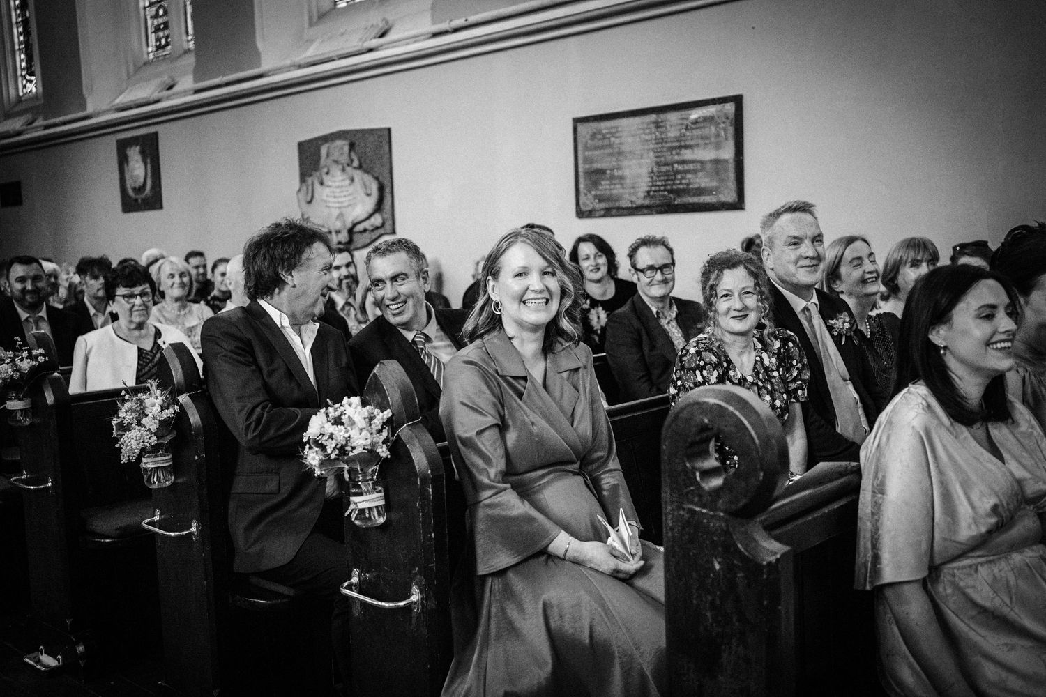 Black and white photo of wedding guests smiling and laughing while seated in church pews during a ceremony.