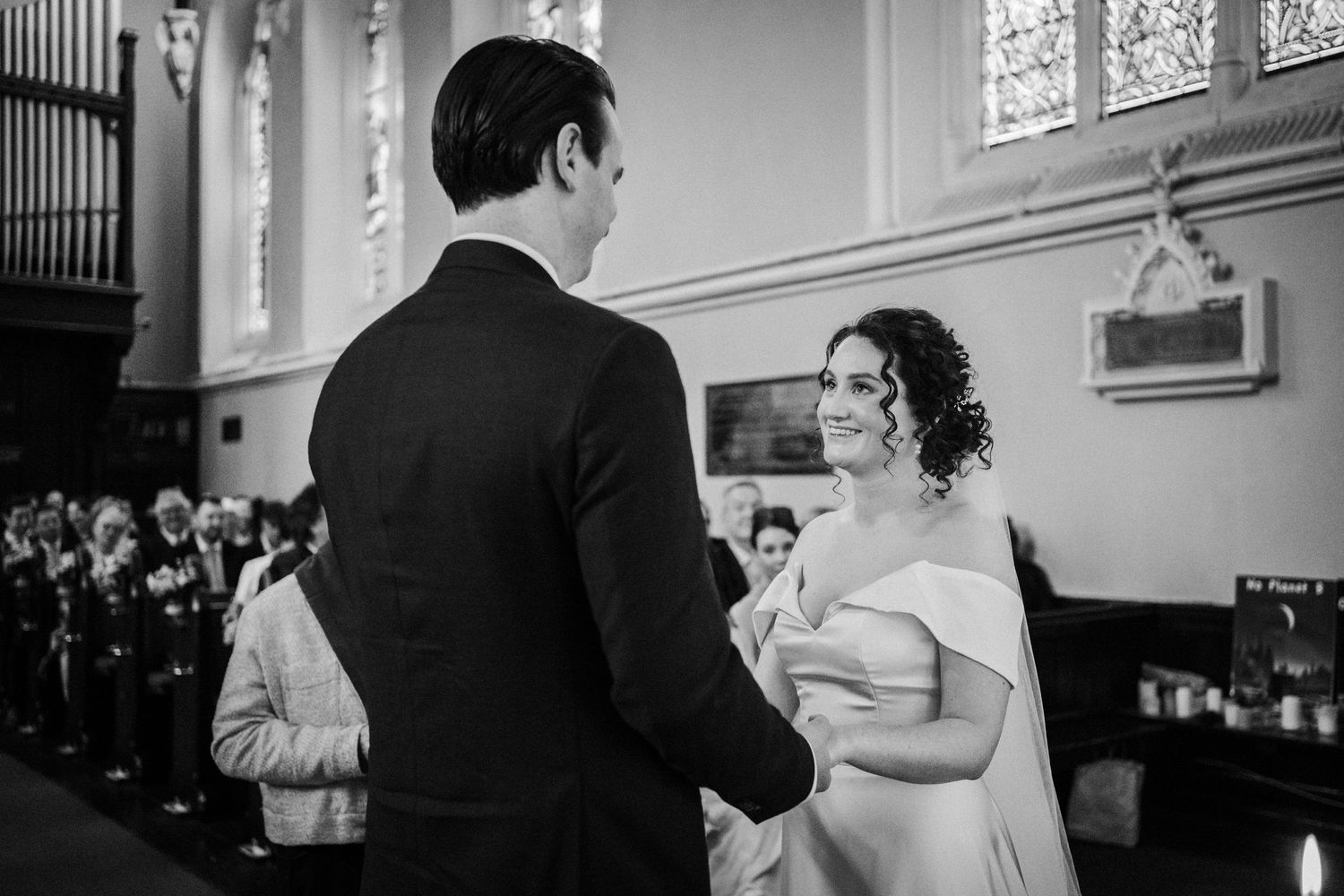 A black and white wedding ceremony photo inside a bright church with stained glass windows and an organ in the background.