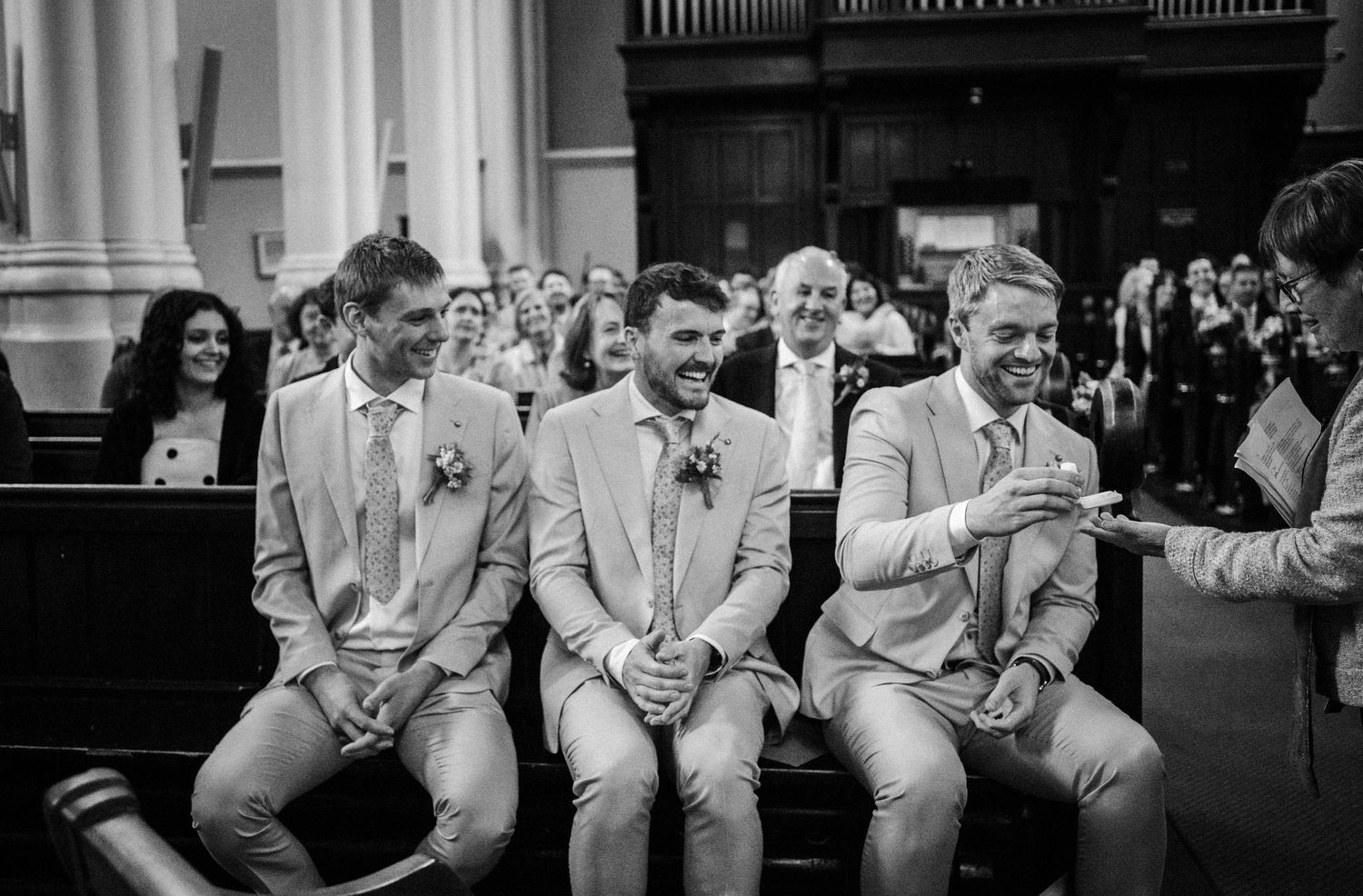 Three wedding party members in light suits sit together on a pew during a black and white church ceremony.