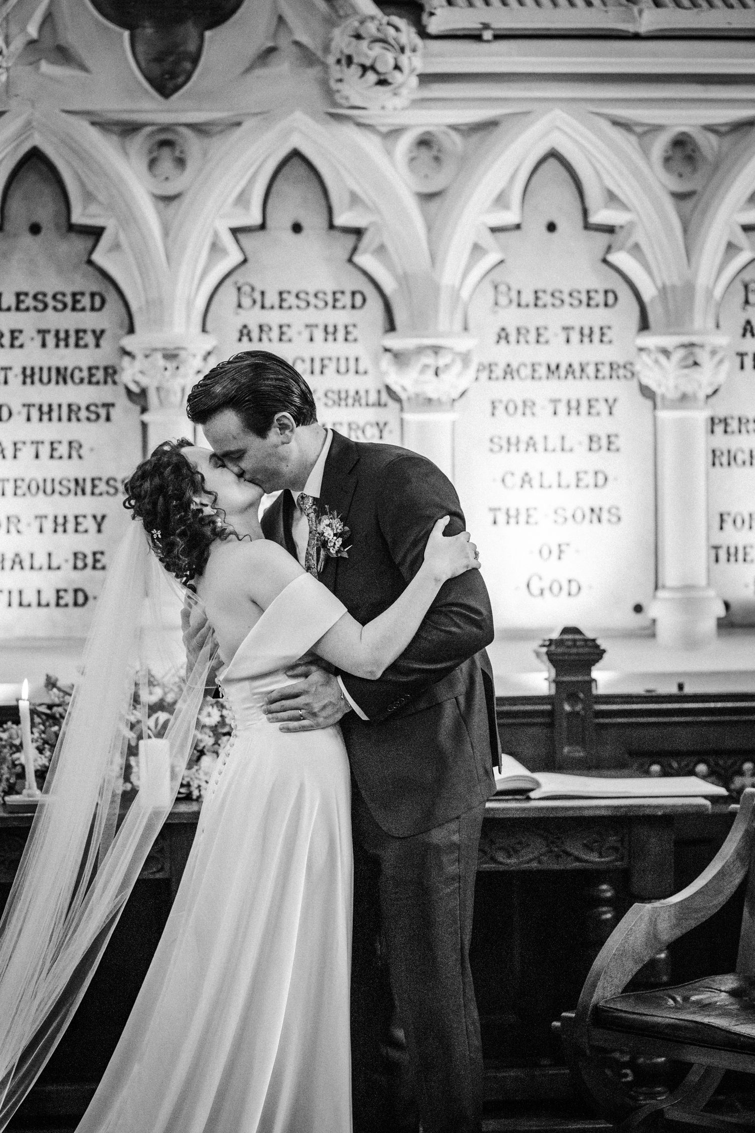 A romantic wedding kiss in a church with gothic arches and biblical text inscribed on the wall behind the couple.