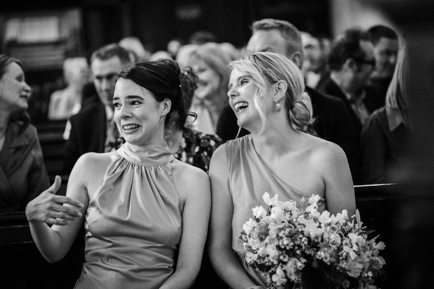 Two elegantly dressed wedding guests share a joyous laugh together while sitting at a black and white ceremony.
