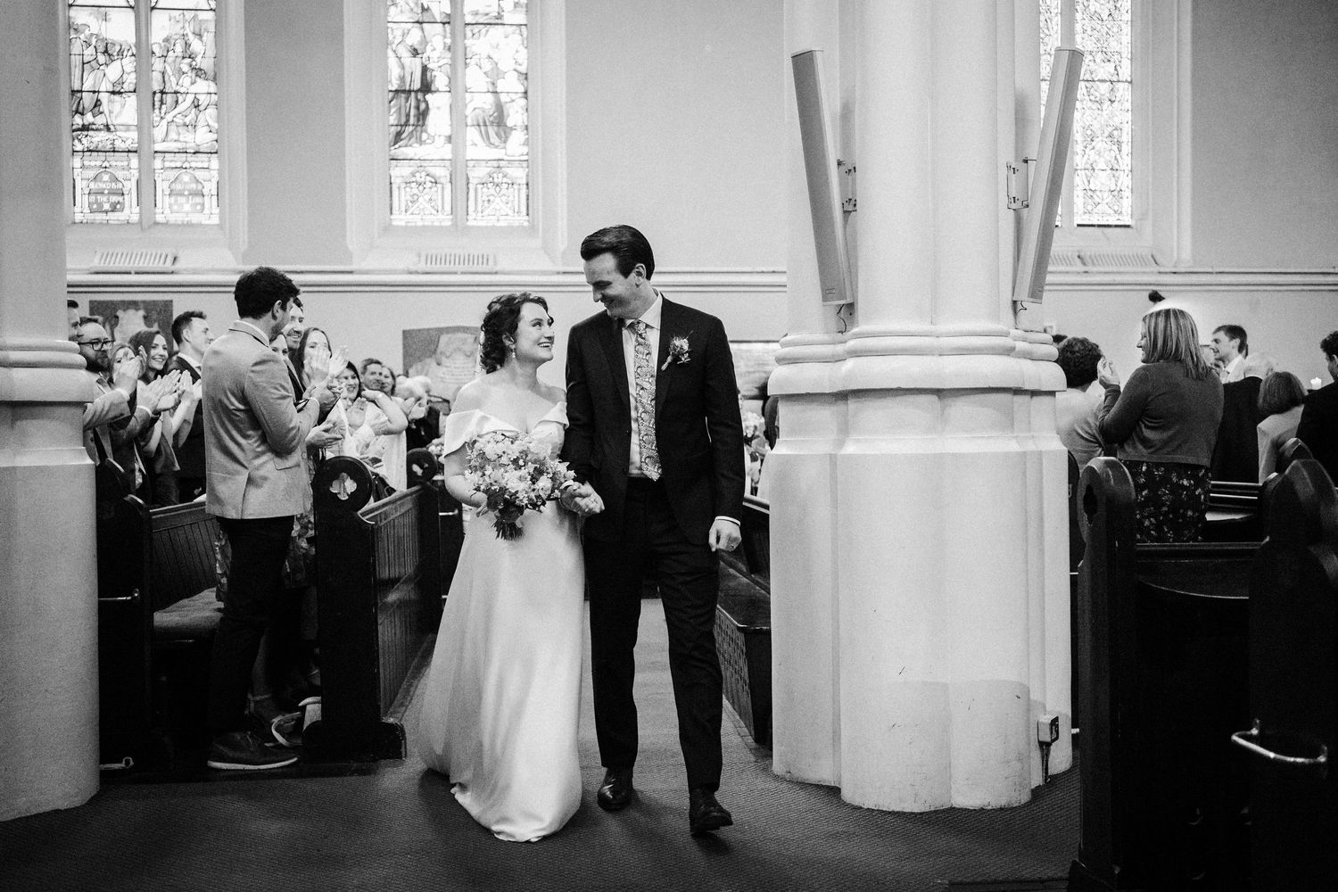 A bride and groom walk down the aisle in a church as guests look on in this black and white candid ceremony photo.