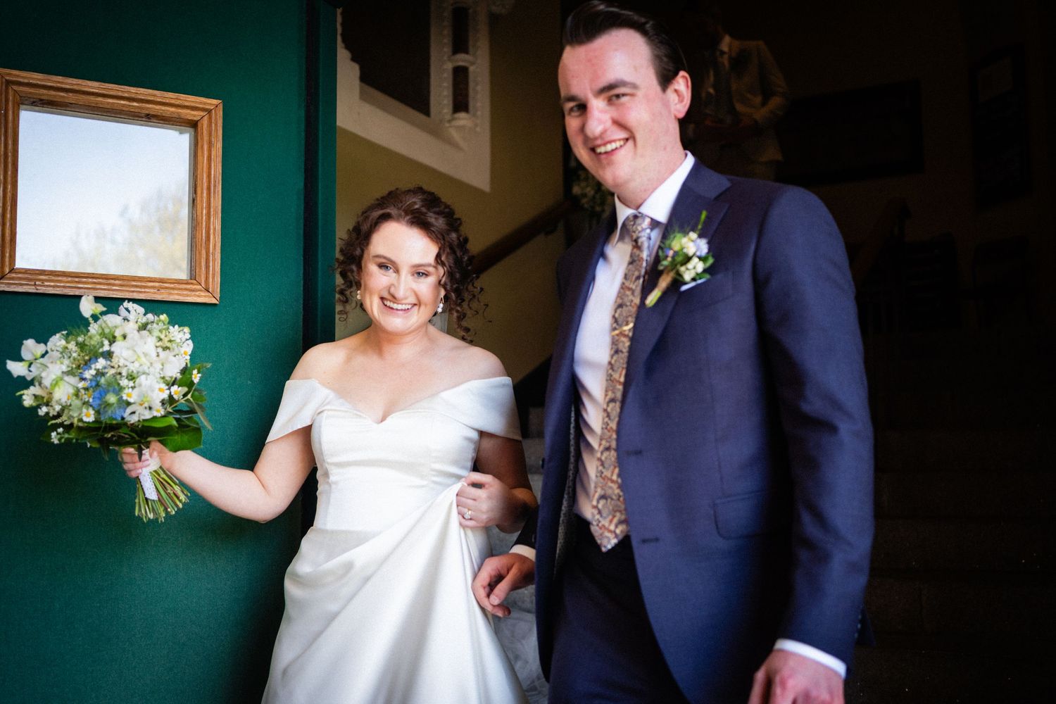 A couple dressed in formal wedding attire stand together against a green wall with floral bouquet.
