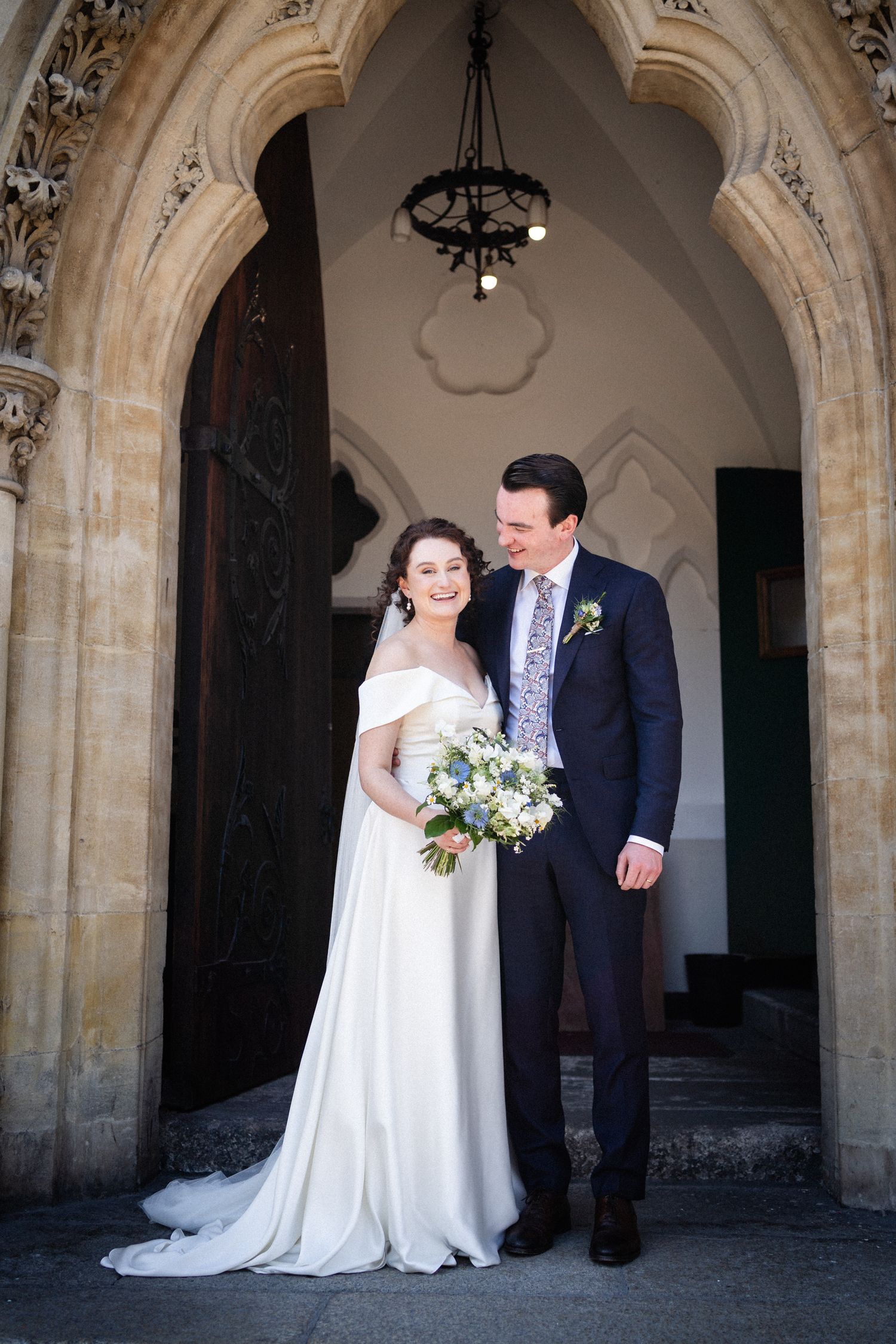 A couple stands in front of a gothic stone archway on their wedding day, with the bride holding a blue and white bouquet.
