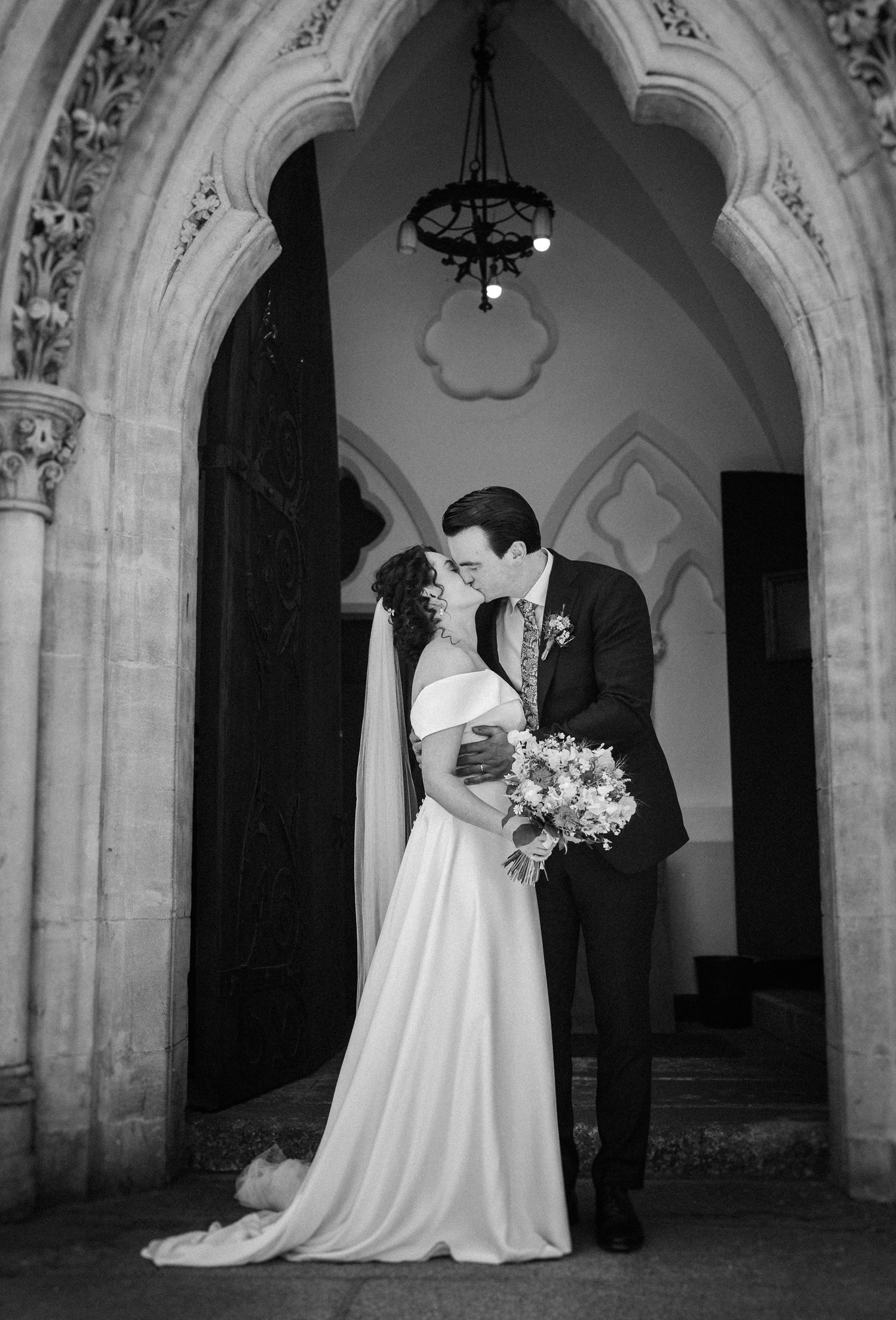 A newlywed couple sharing an intimate moment under a gothic archway in elegant black and white photography.