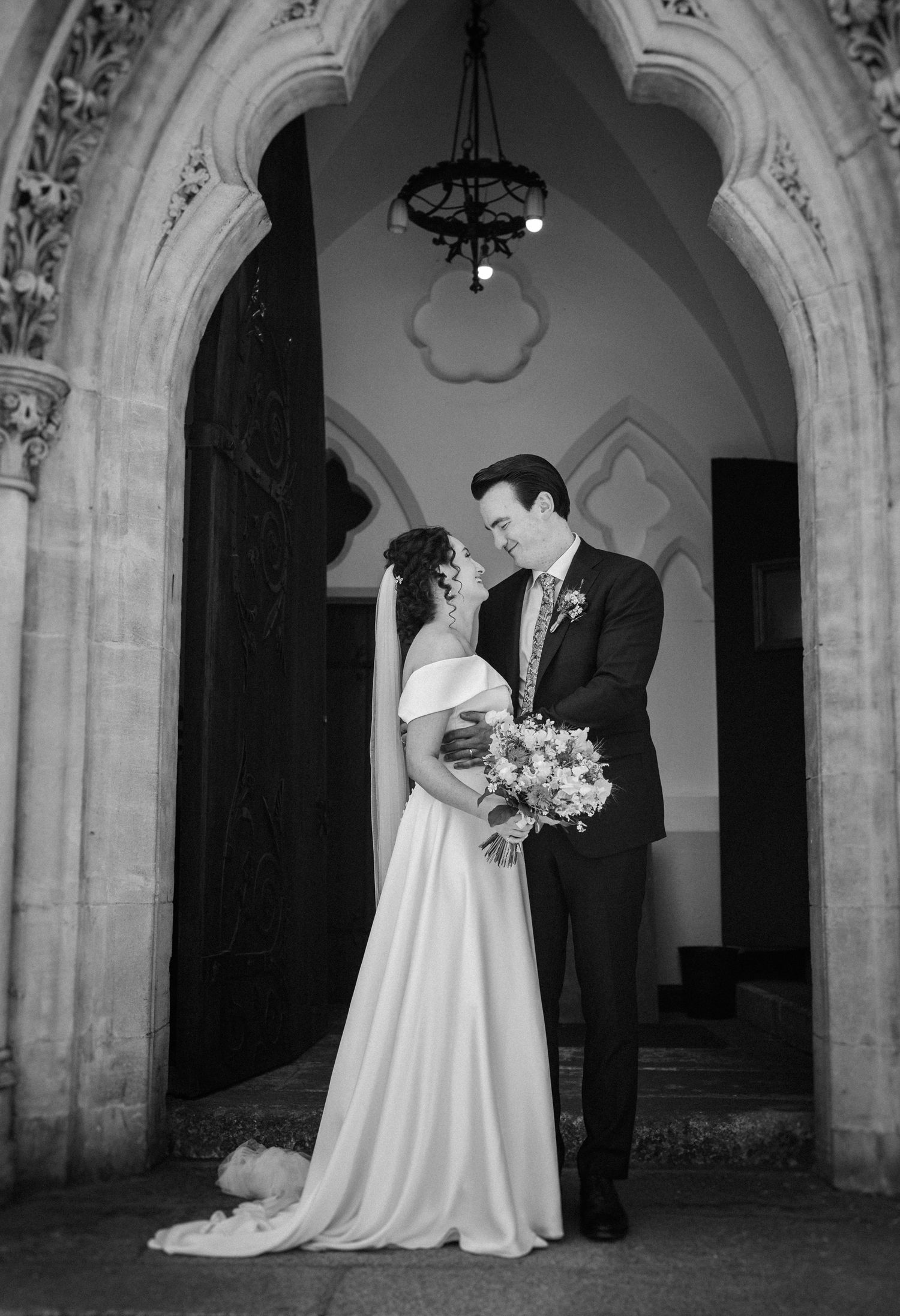 Black and white wedding photo of a couple standing in a gothic stone archway with ornate details and hanging chandelier.