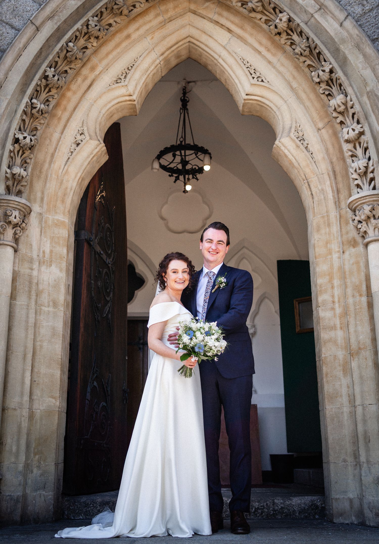 A couple stands under an ornate gothic stone archway entrance with decorative carving and chandelier at their wedding ceremony.