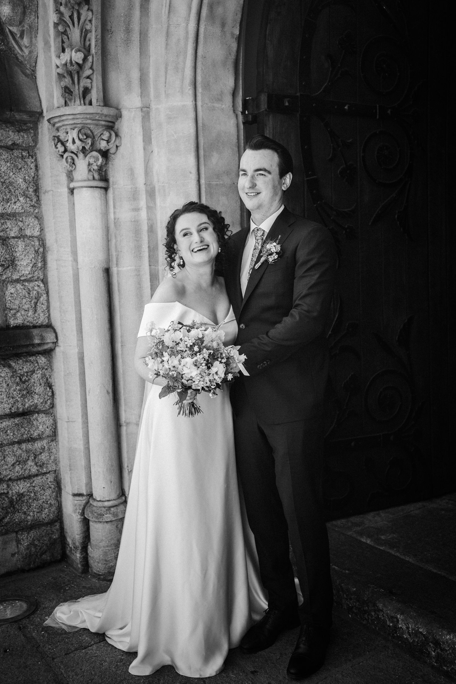 Black and white wedding portrait of a couple standing by ornate stone columns at a church entrance.