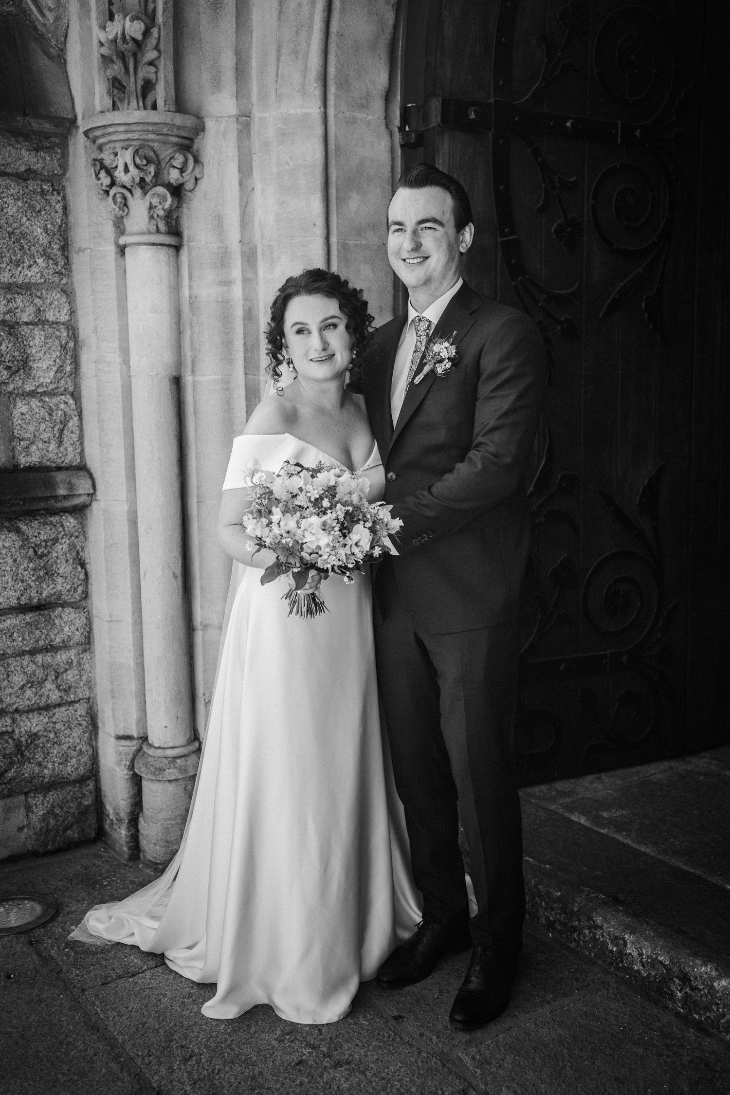 Black and white wedding photo of newlyweds posing together beside decorative church architecture.