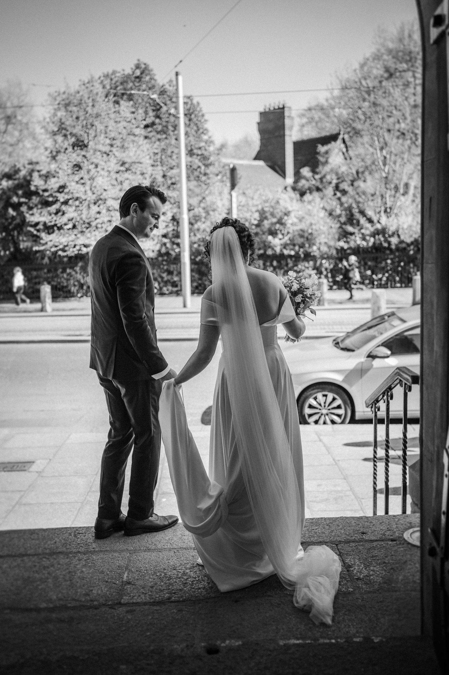 A bride in a flowing white gown and her groom stand together at the entrance steps on their wedding day.