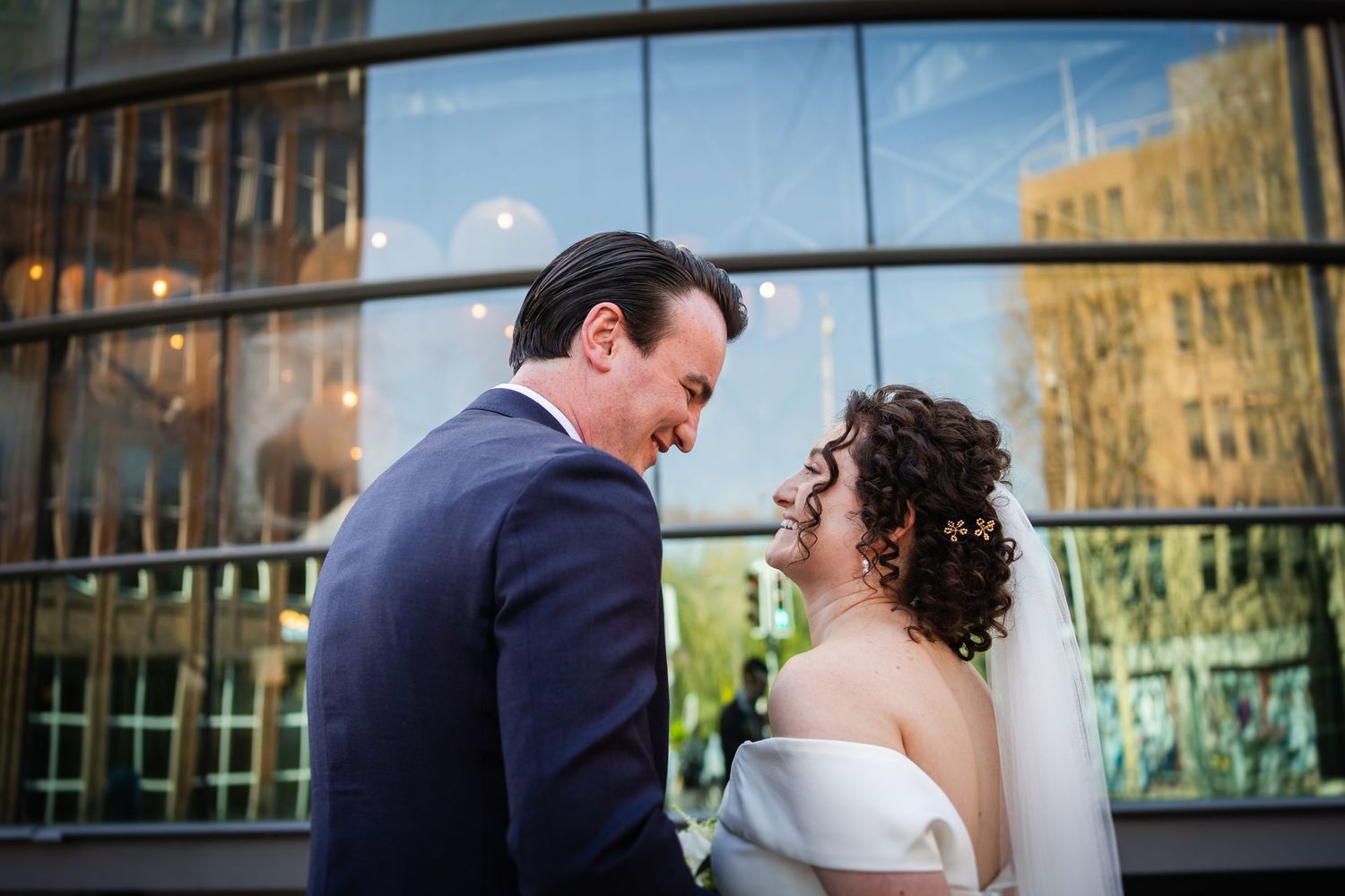 A sweet wedding moment shared between newlyweds in front of a glass building reflecting the urban cityscape.