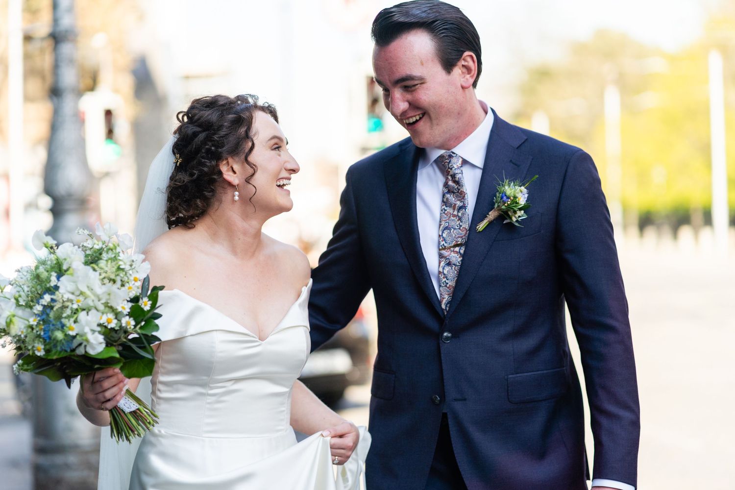 Happy couple in wedding attire walks outdoors sharing a joyful moment with white floral bouquet.