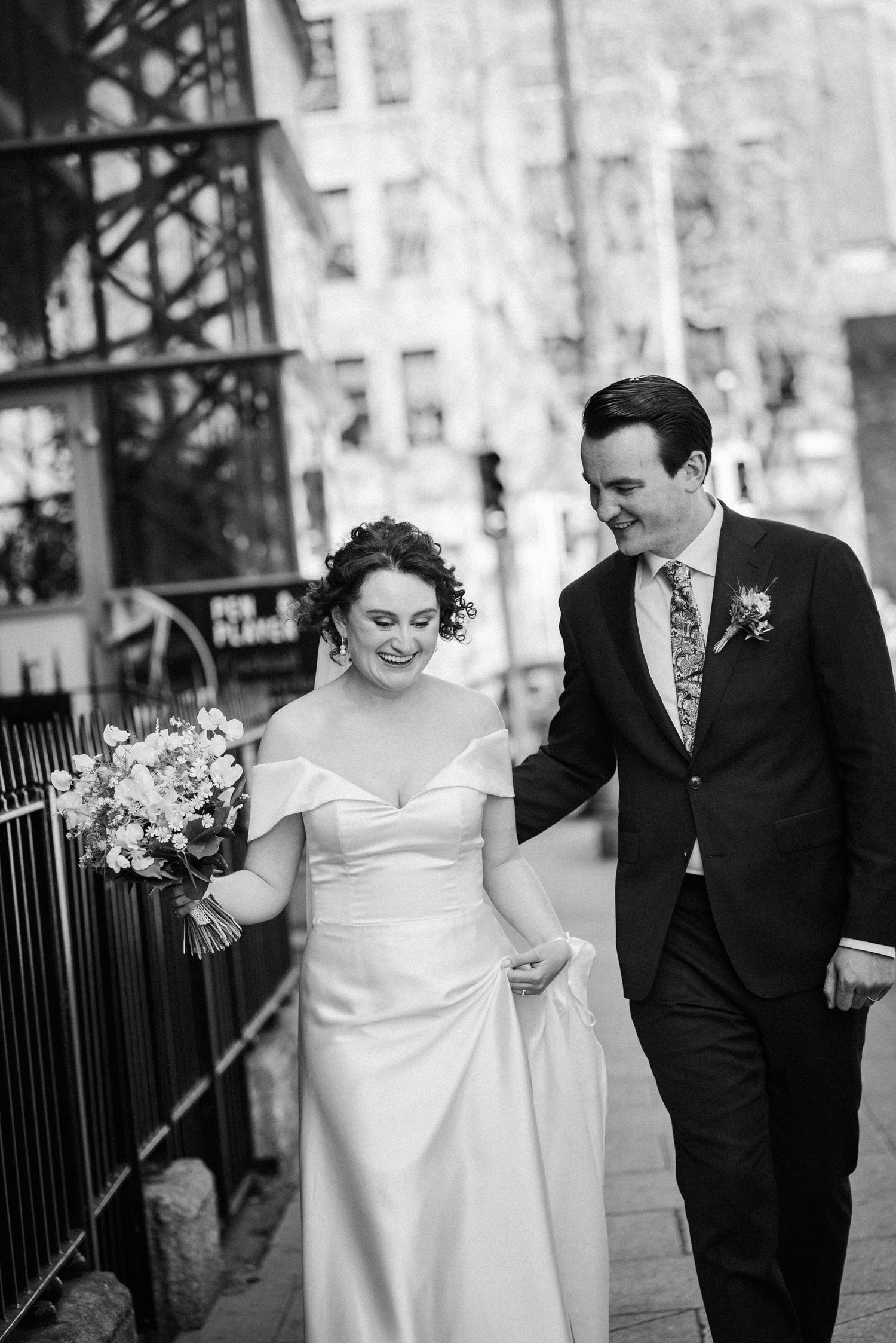 Black and white photo of newlyweds walking together on city street, bride in sleek dress holding floral bouquet.