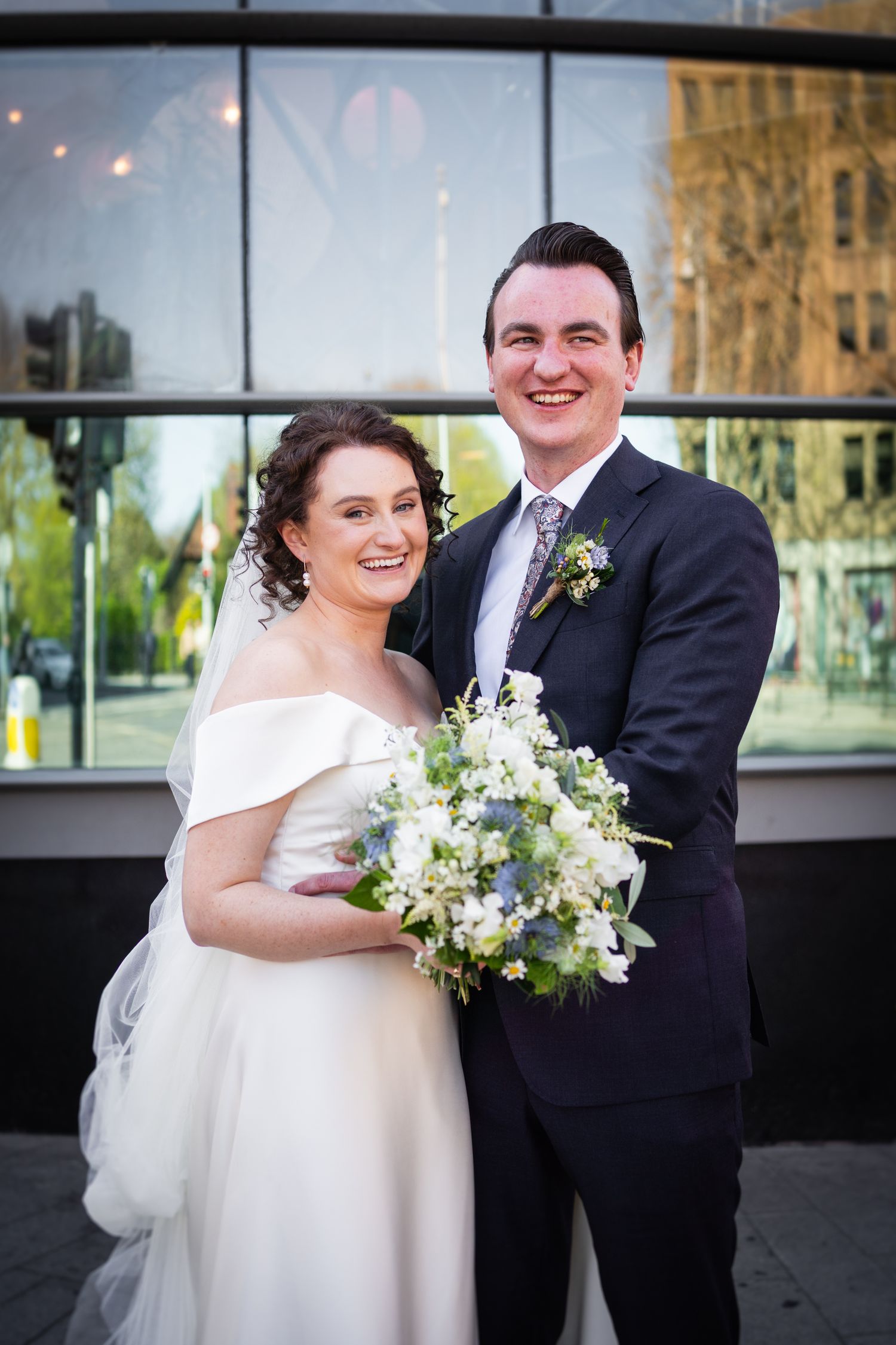 A bride in an off-shoulder wedding gown holds a white floral bouquet while posing with a groom in a dark suit outdoors.