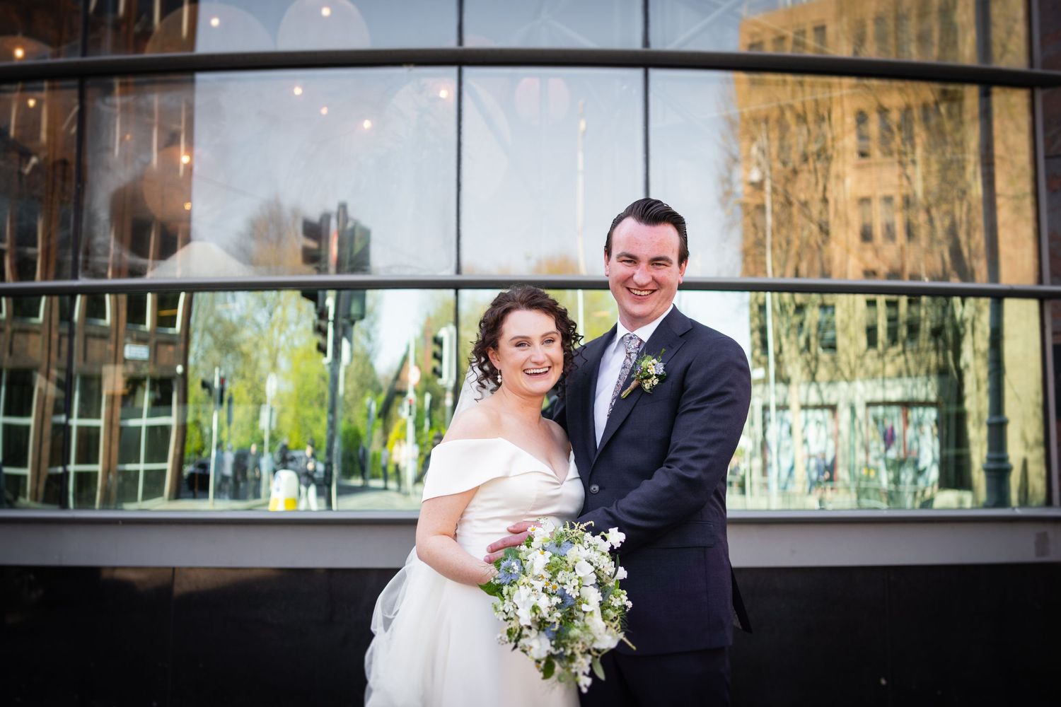A happy couple in wedding attire stands together smiling in front of a modern glass building exterior.
