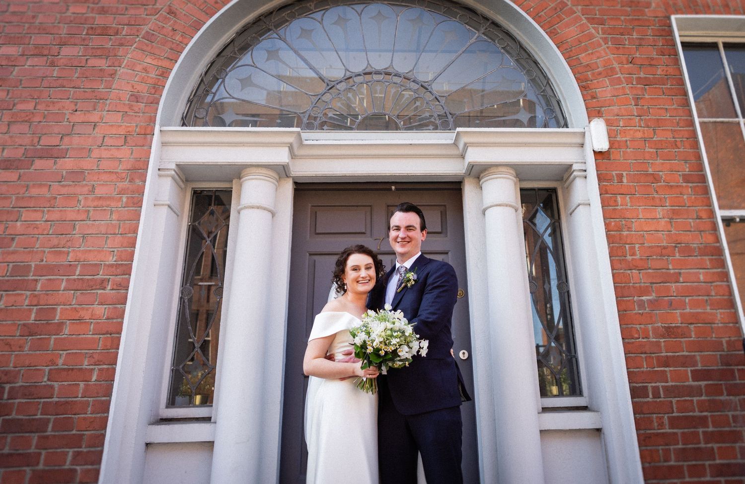 A couple stands in front of a classic red brick building entrance with white columns and an arched doorway.