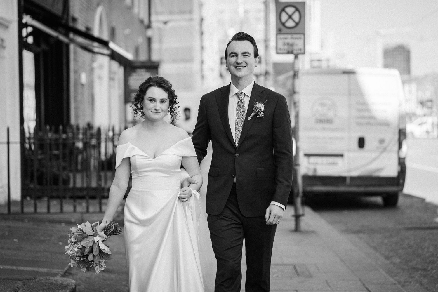 A couple in wedding attire walking together on a city street in black and white photography.