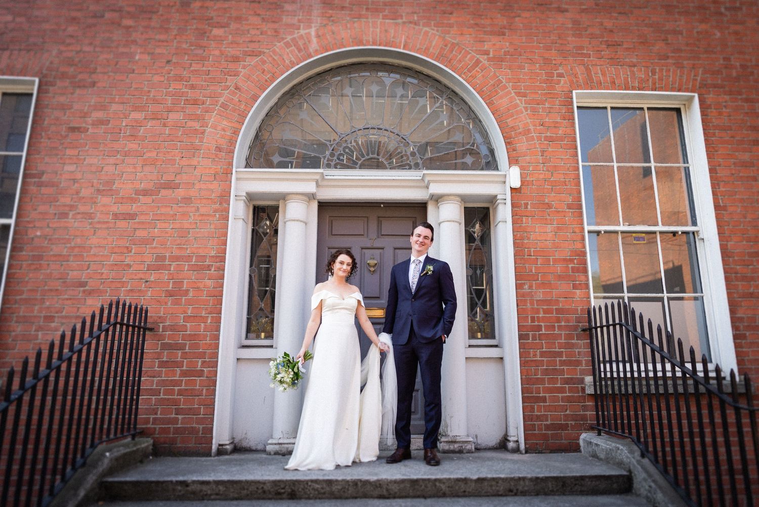 A couple stands in a wedding dress and suit at a grand brick building entrance with white columns and an arched doorway.