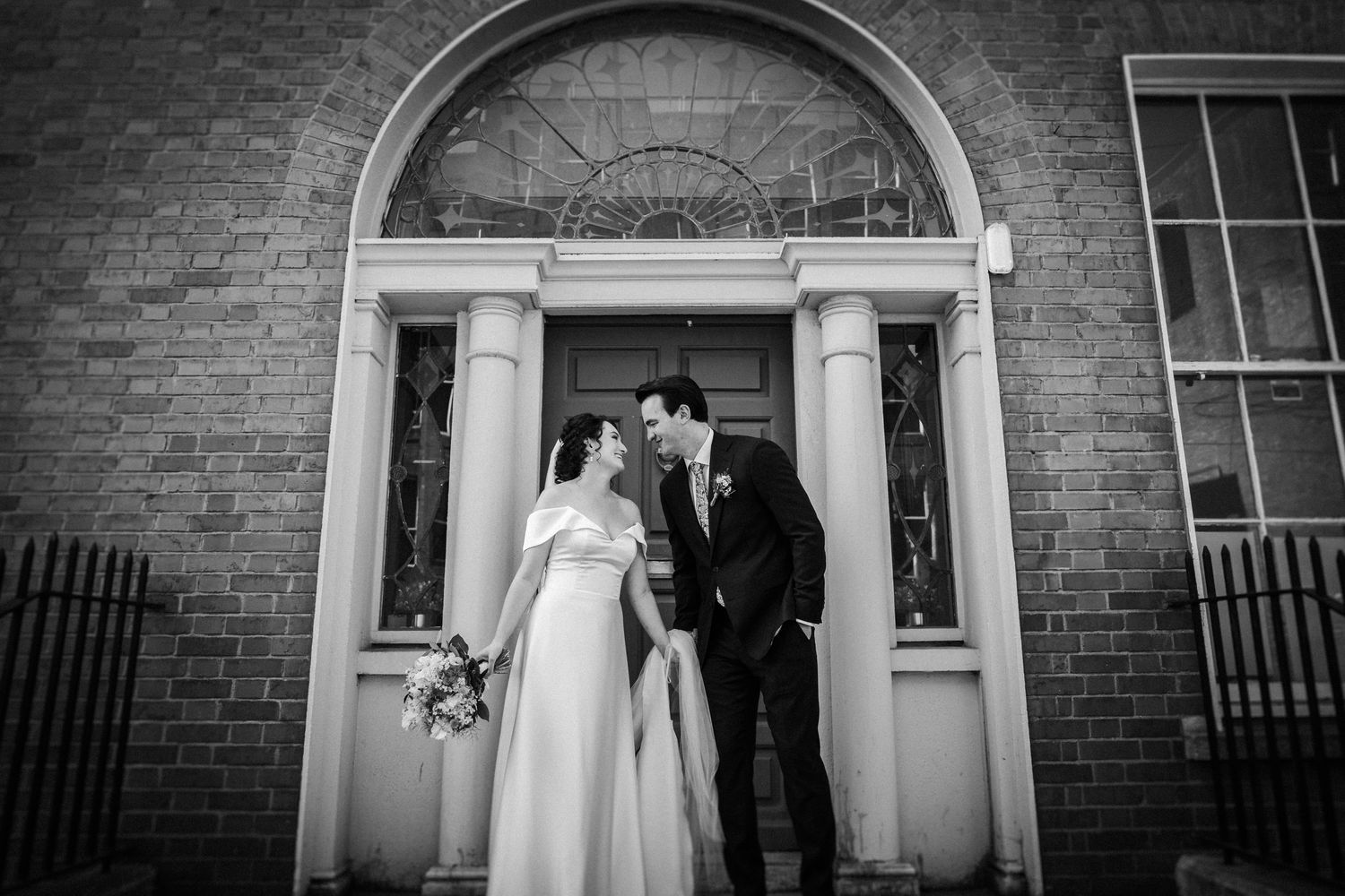 A black and white photograph of a couple standing in front of an elegant Georgian doorway with arched transom window.