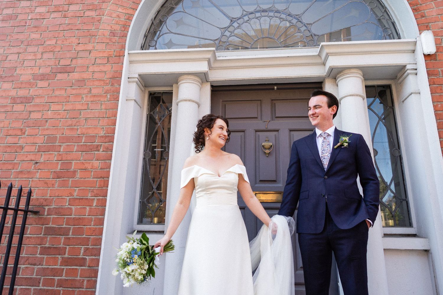 A couple stands smiling in front of an elegant Georgian door with a fanlight window and red brick facade.