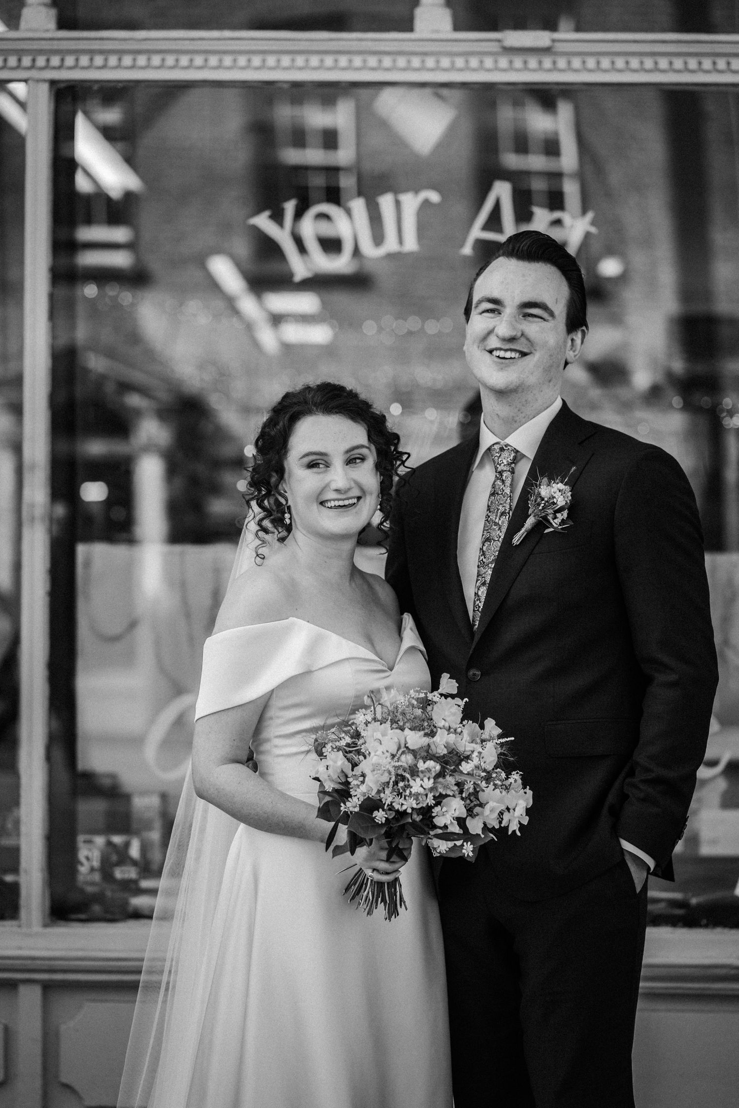 Black and white wedding portrait showing a bride in off-shoulder dress with bouquet and groom in suit outside storefront.