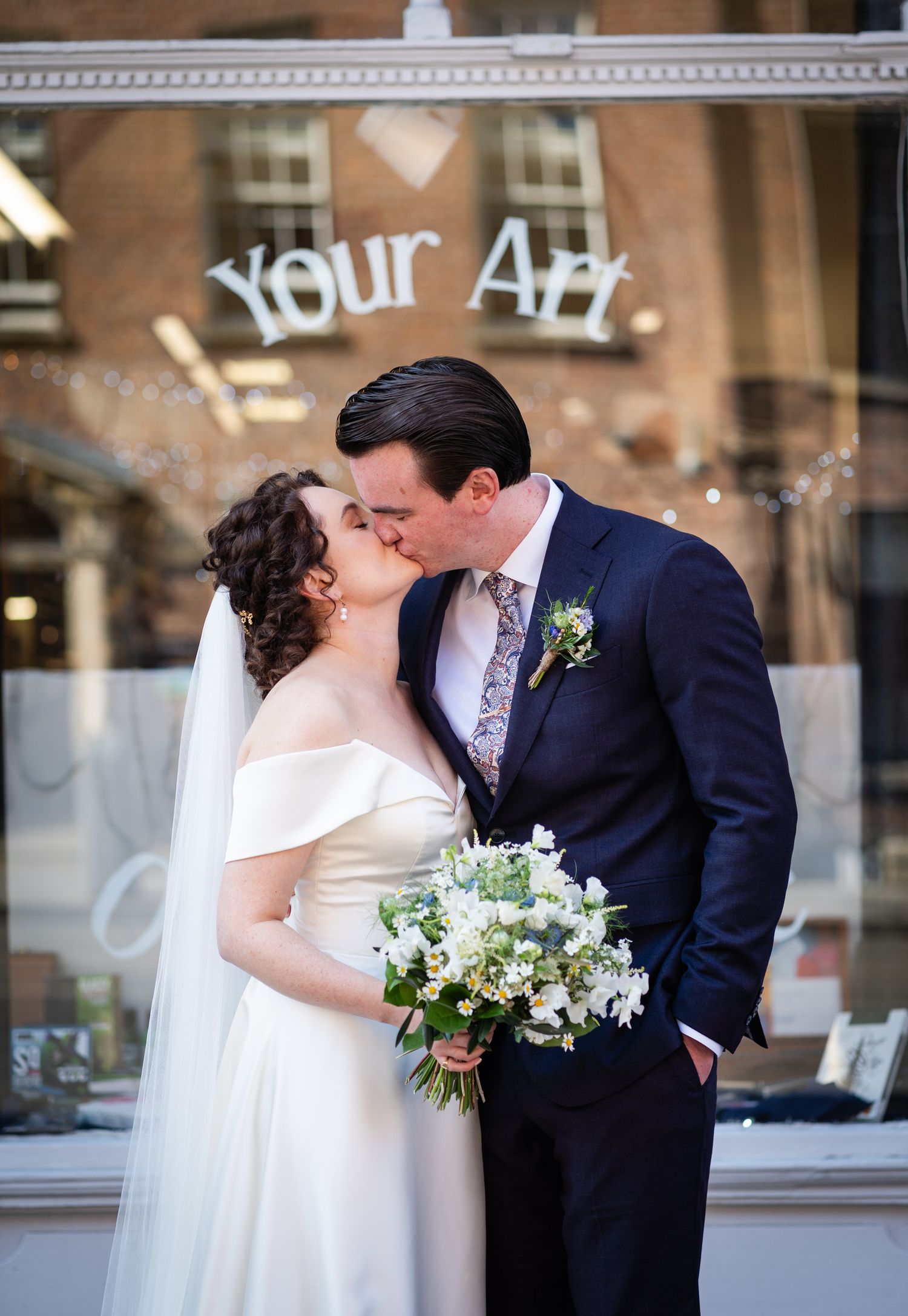 Newlyweds share a romantic kiss in front of storefront window while bride holds white floral bouquet.