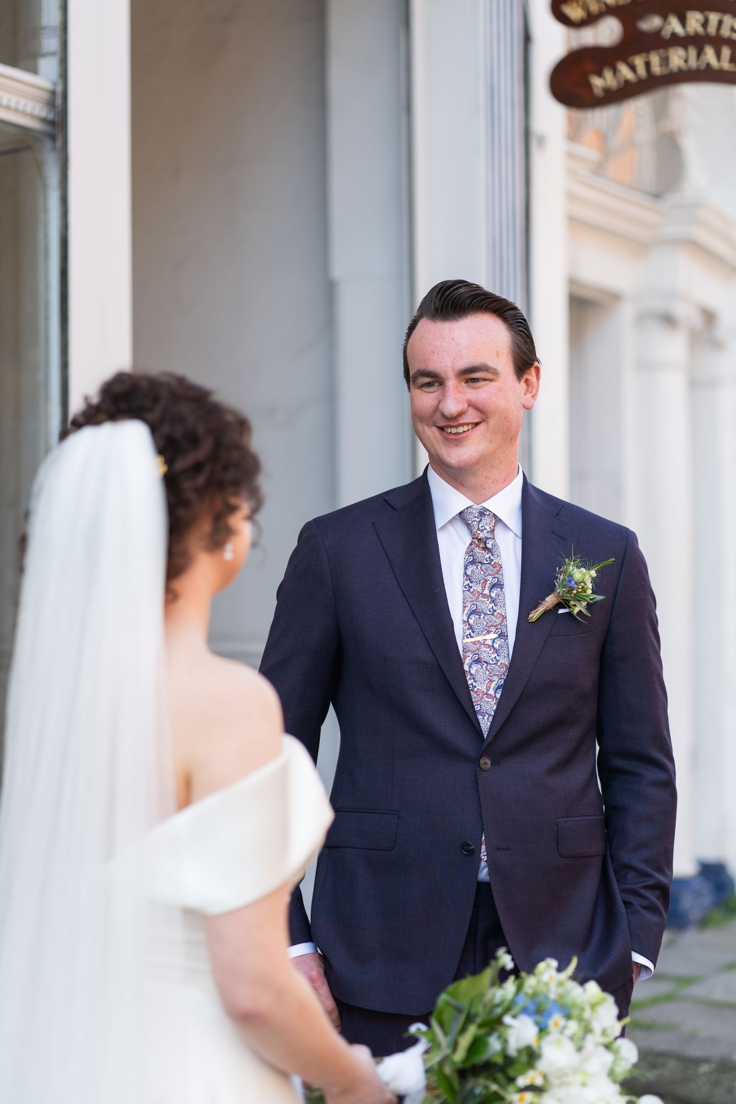 A groom in a navy suit with floral tie and boutonniere smiles at his bride holding a white and blue bouquet.