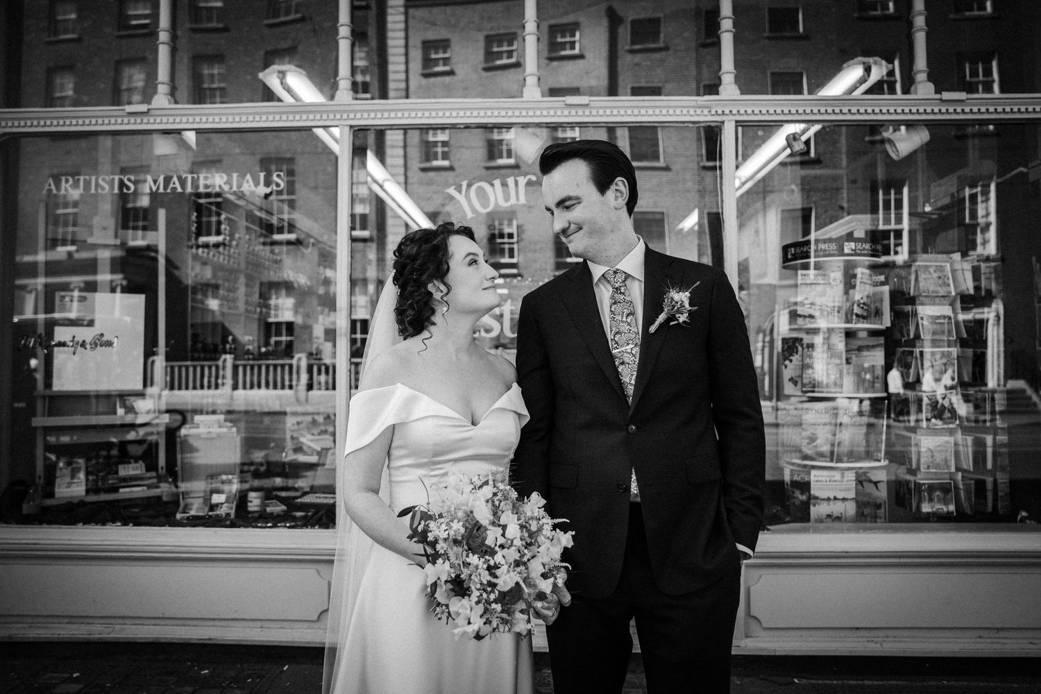 A black and white wedding photo of a couple sharing a joyful moment in front of a building with reflective windows.