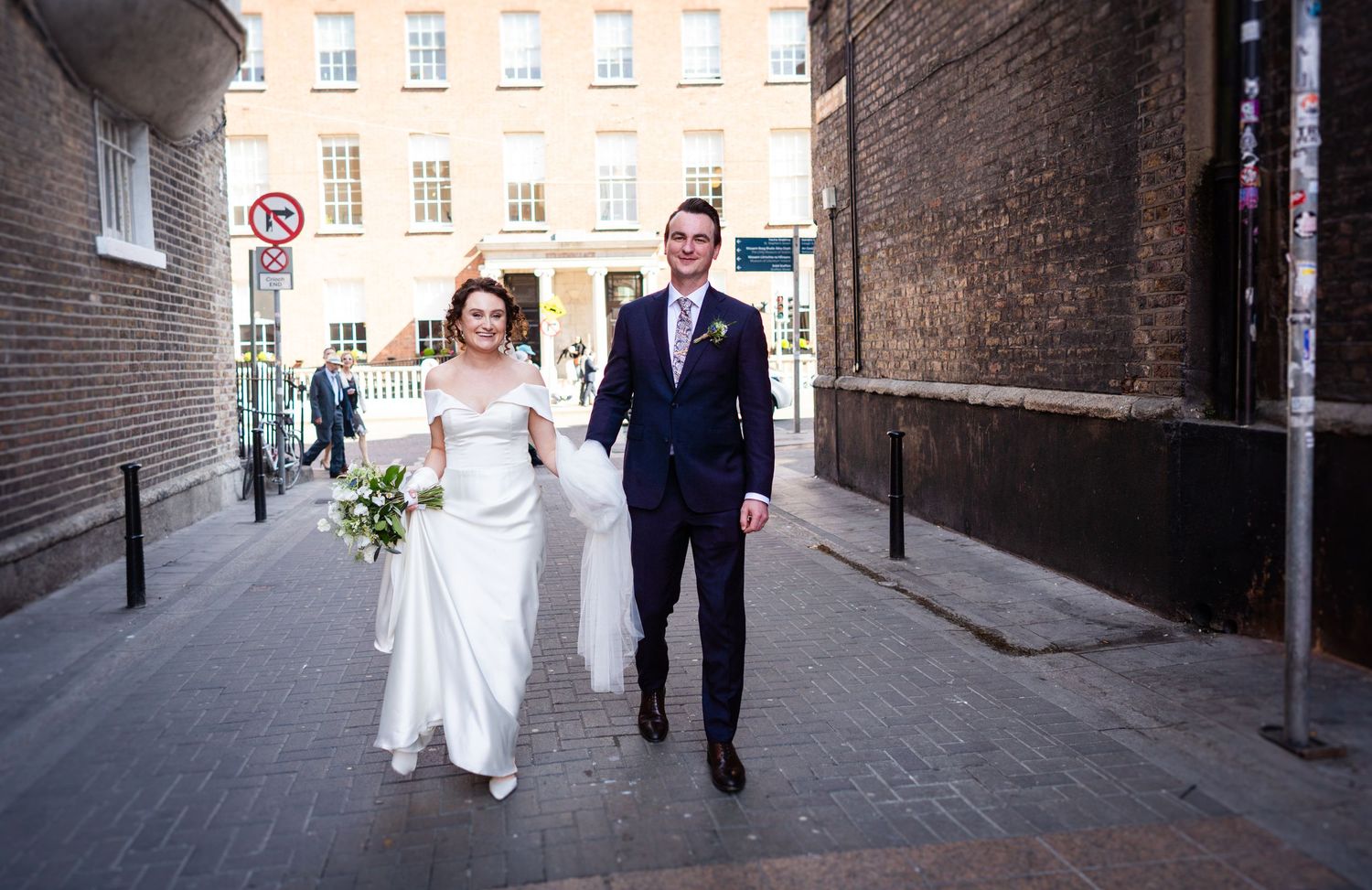 Newlyweds walk together through a historic brick alleyway in formal wedding attire with bouquet.