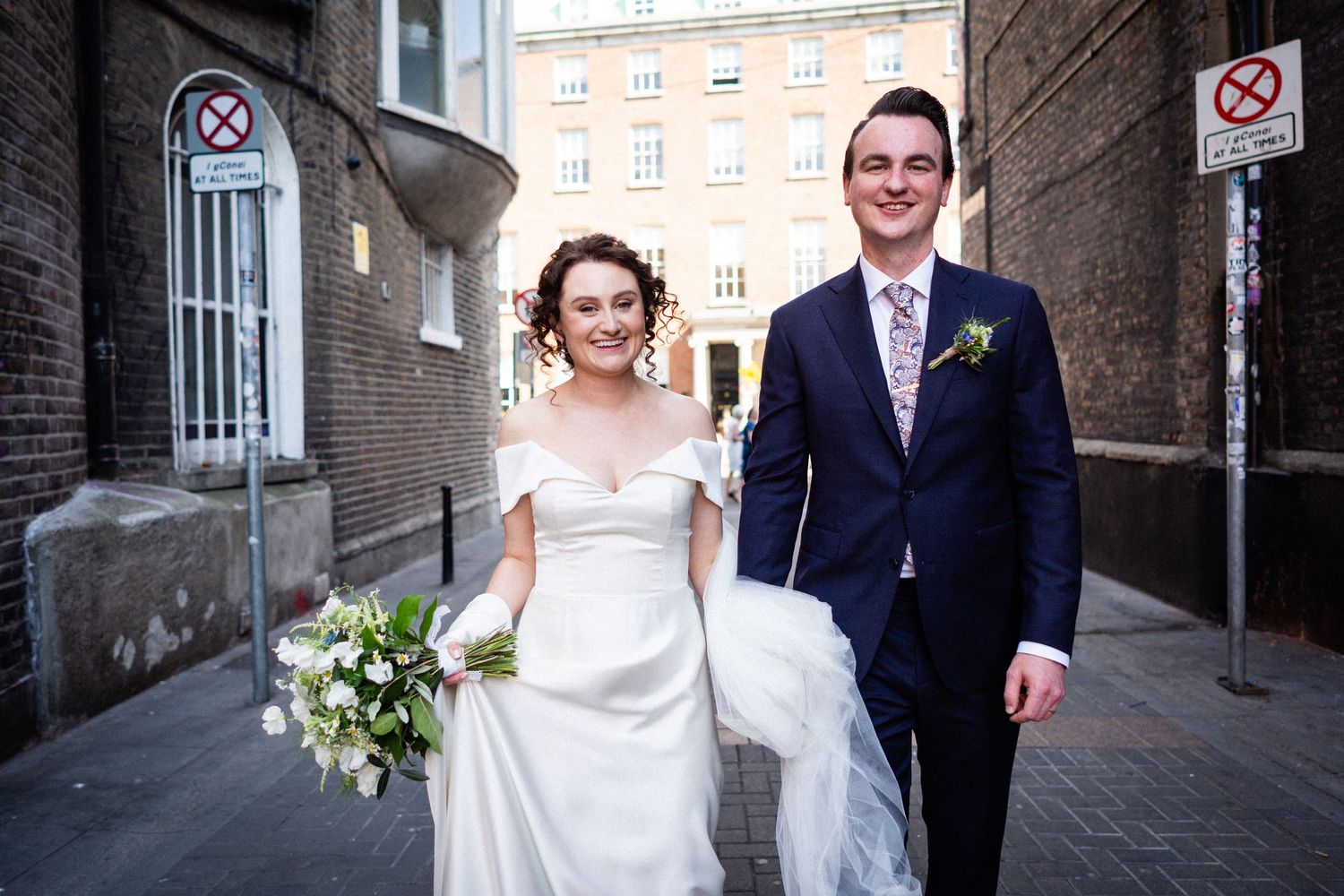 Happy newlywed couple walks together on city street with bride holding white bouquet and wearing elegant dress.