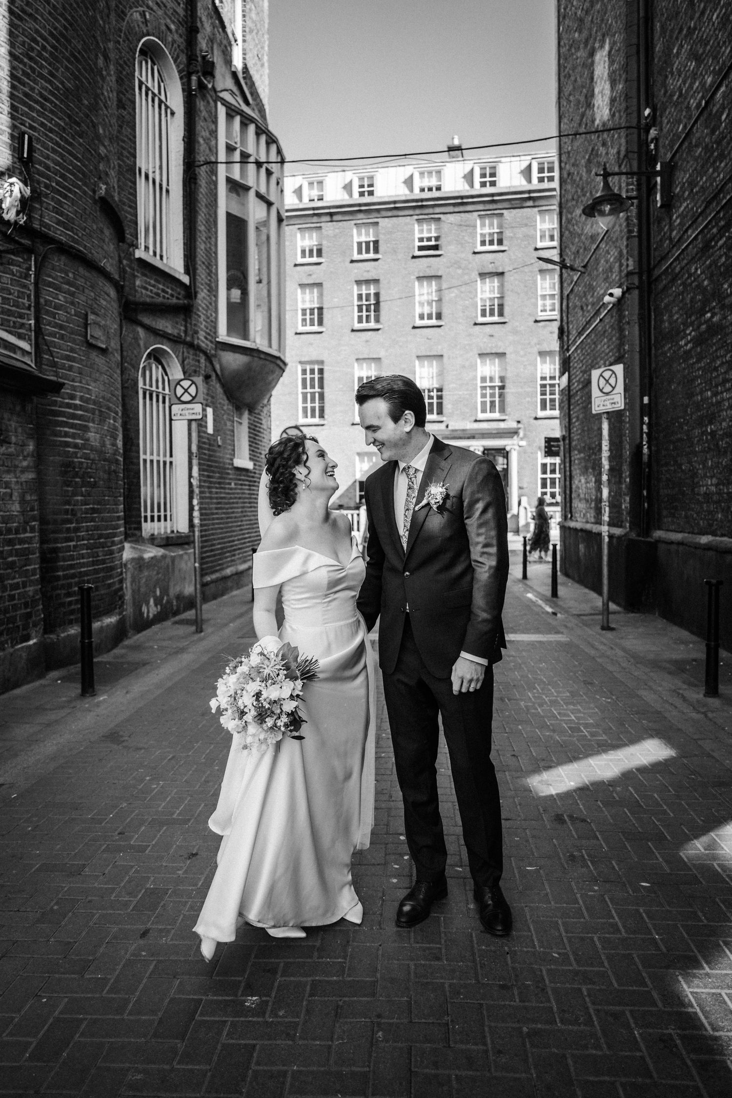 A romantic wedding moment with a couple in elegant attire between brick buildings on a narrow city street.