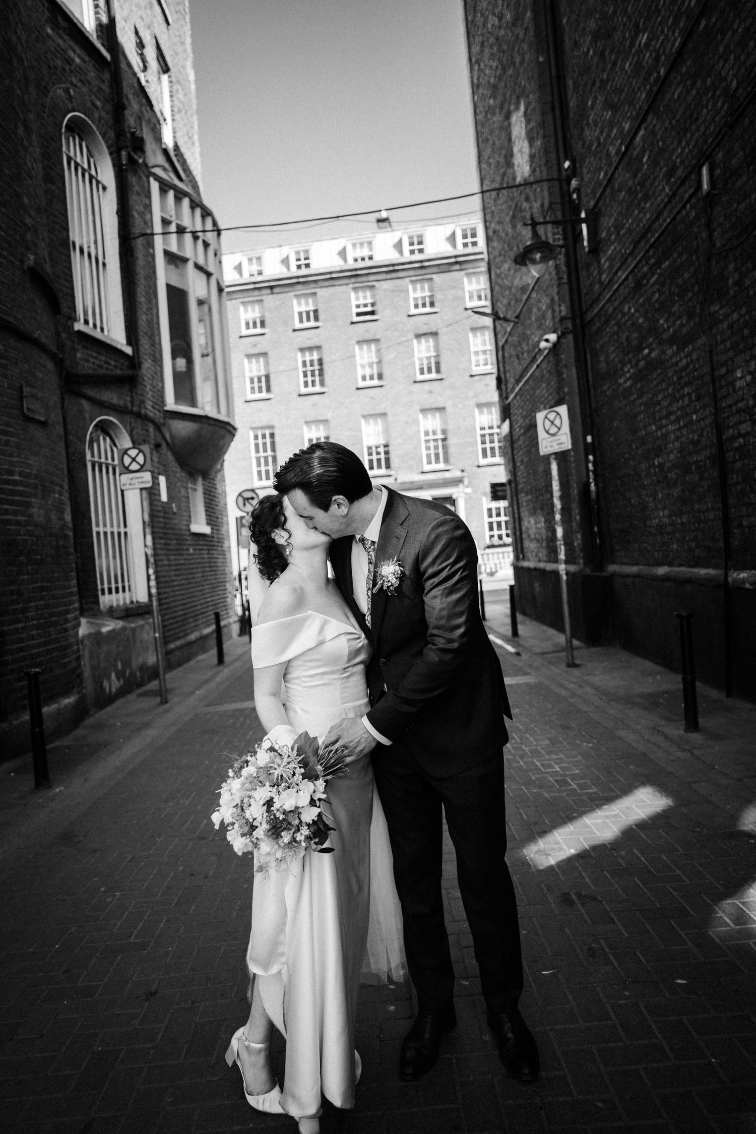 A romantic black and white wedding photograph showing a couple embracing in an urban alley with brick buildings.
