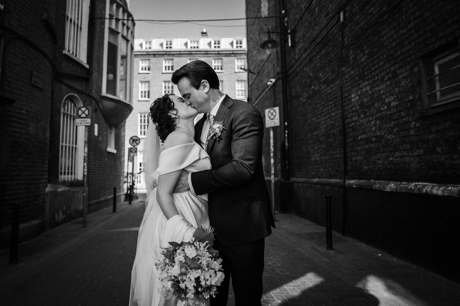 Elegant newlywed couple shares intimate moment in urban alley between brick buildings with floral bouquet.