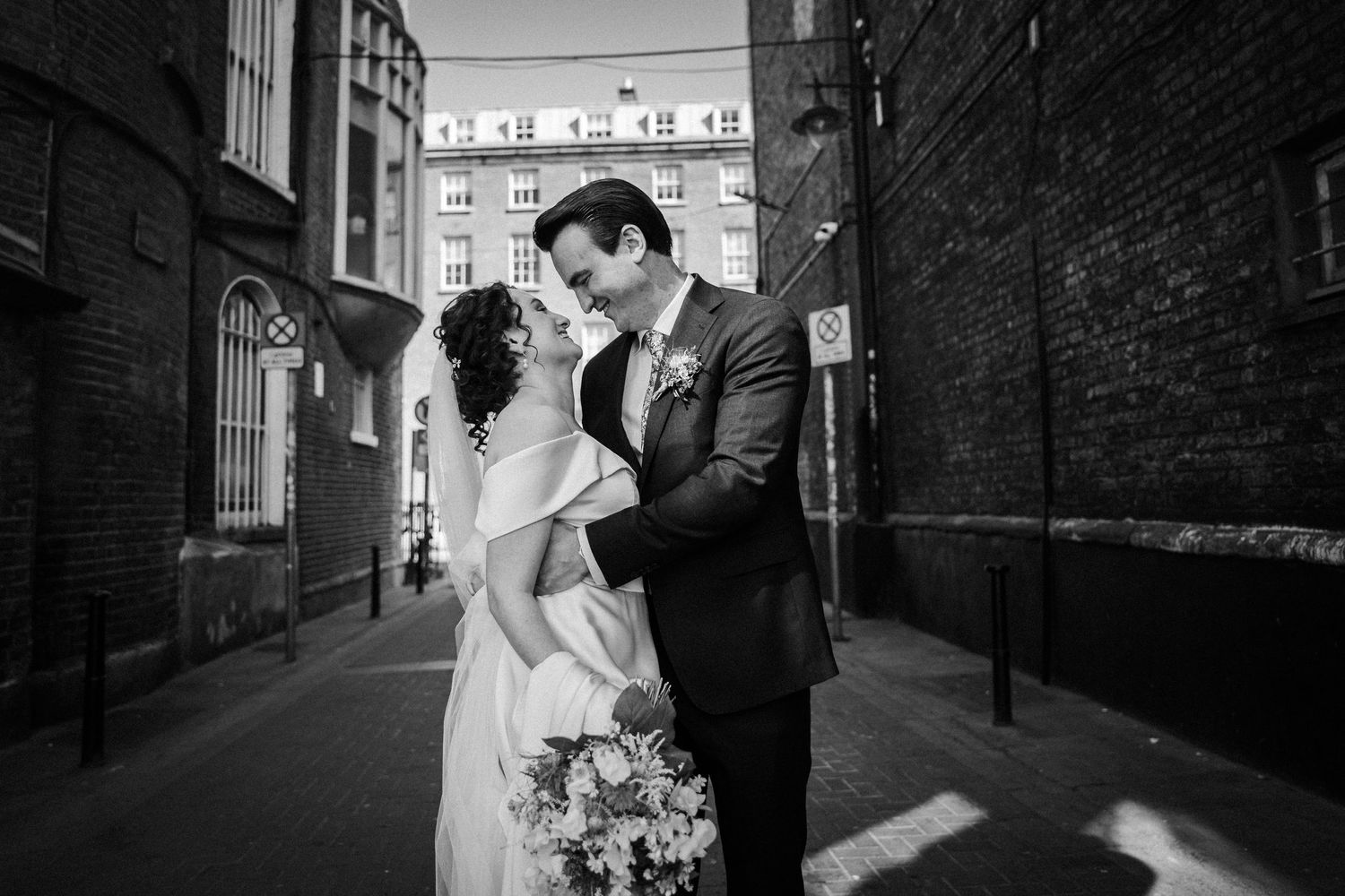Black and white wedding photograph of a couple embracing in an urban alleyway with brick walls and bouquet.
