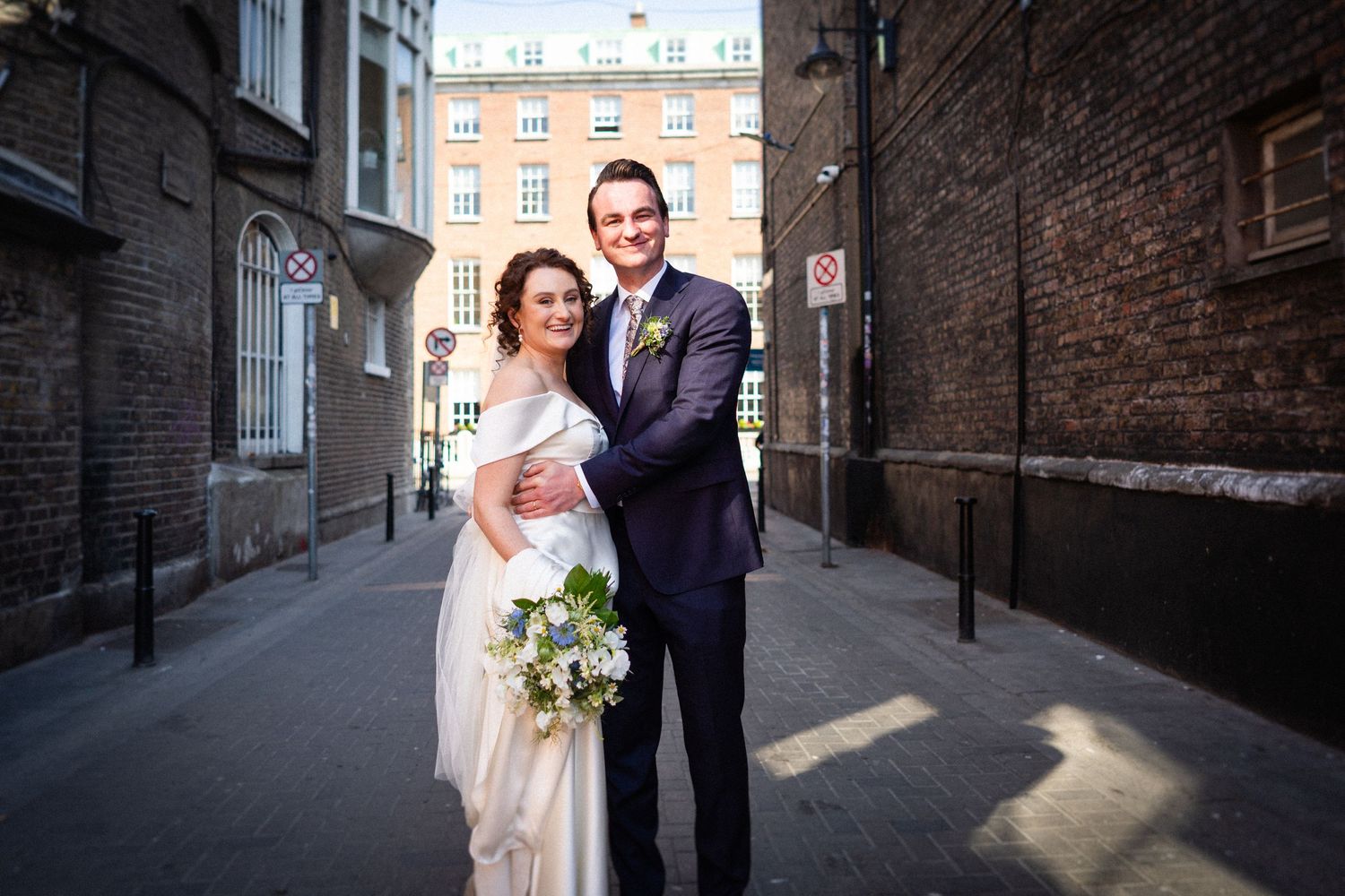A wedding couple poses in a charming brick alleyway with sunlit shadows and historic architecture behind them.