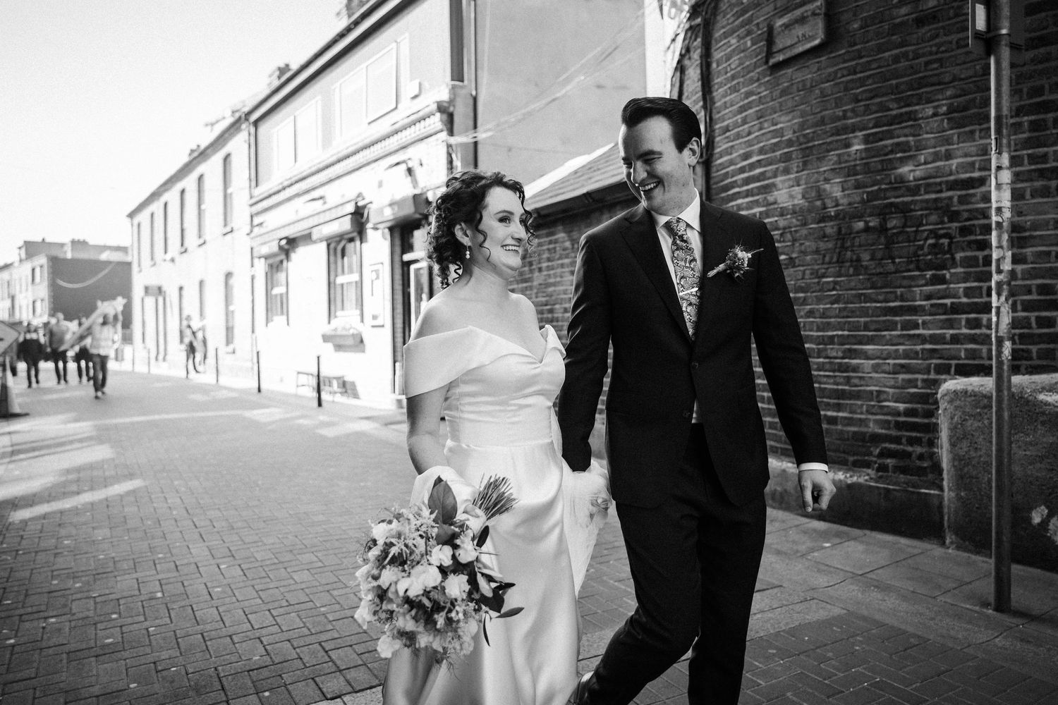 Black and white photo of a newlywed couple walking together on a brick sidewalk, with the bride holding a floral bouquet.