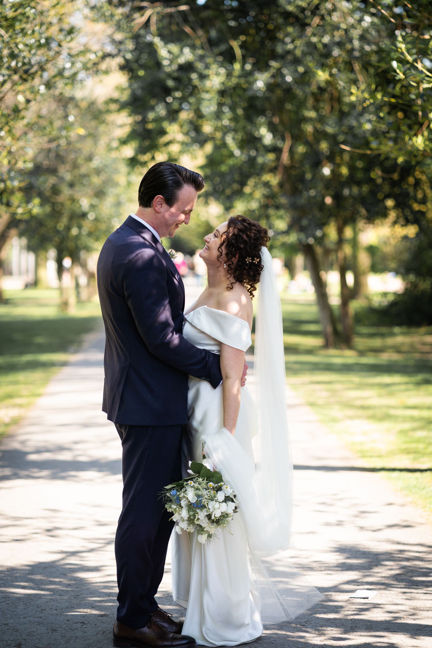 A romantic wedding couple embraces on a tree-lined path while holding a white floral bouquet.