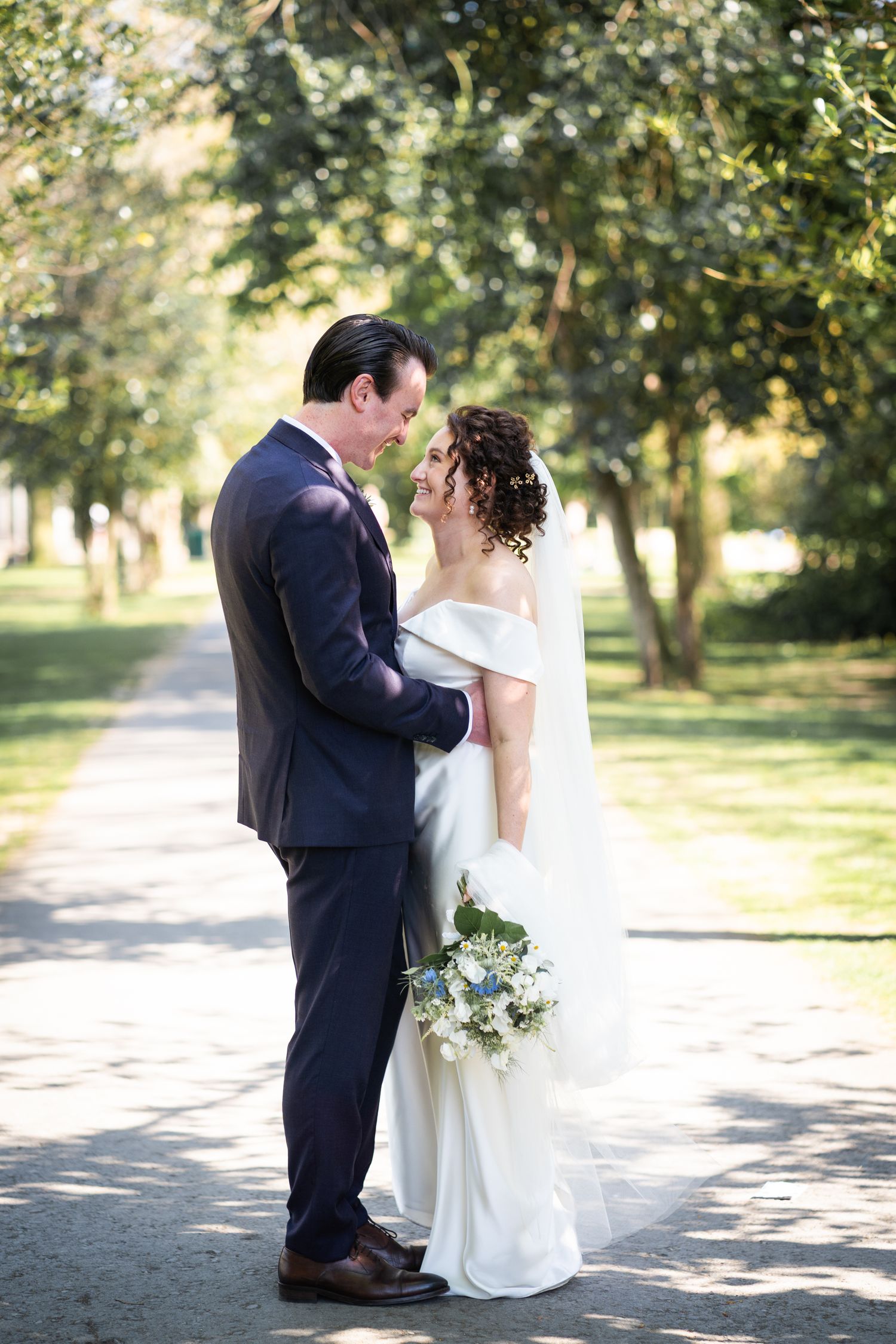 A romantic wedding couple sharing an intimate moment on a tree-lined path outdoors, with a white bridal bouquet.
