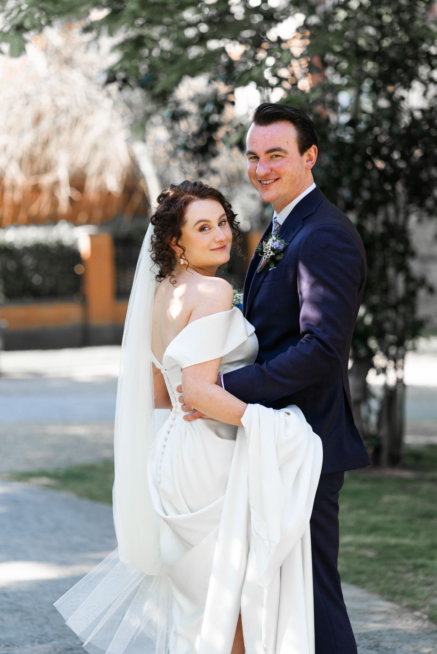 A romantic wedding portrait outdoors showing a couple embracing against a soft, garden-like backdrop with natural lighting.