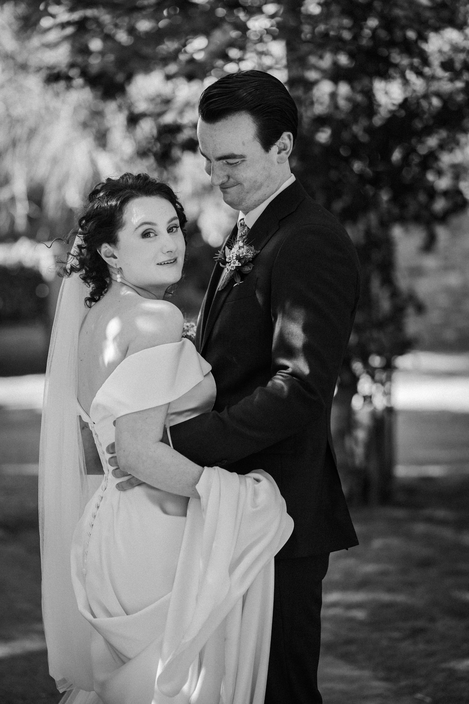 A black and white wedding photograph capturing a romantic moment between a bride in white and groom in a dark suit outdoors.
